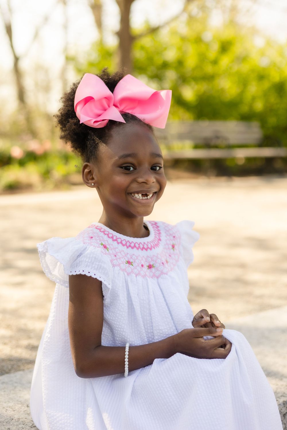 A child in white dress with pink bow smiles brightly during an outdoor photo session.
