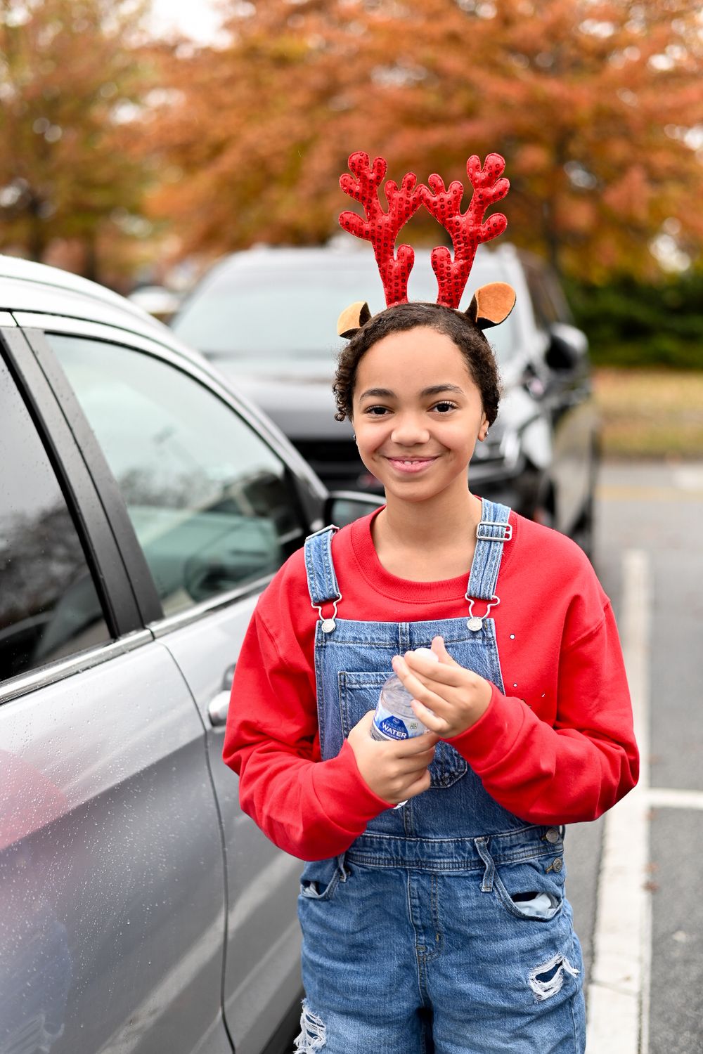 Person in red sweater and overalls wearing festive reindeer antlers in parking lot.