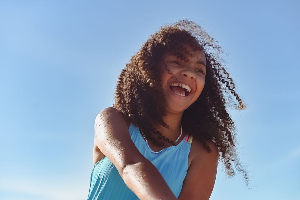 Person with curly hair laughing joyfully against bright blue sky.