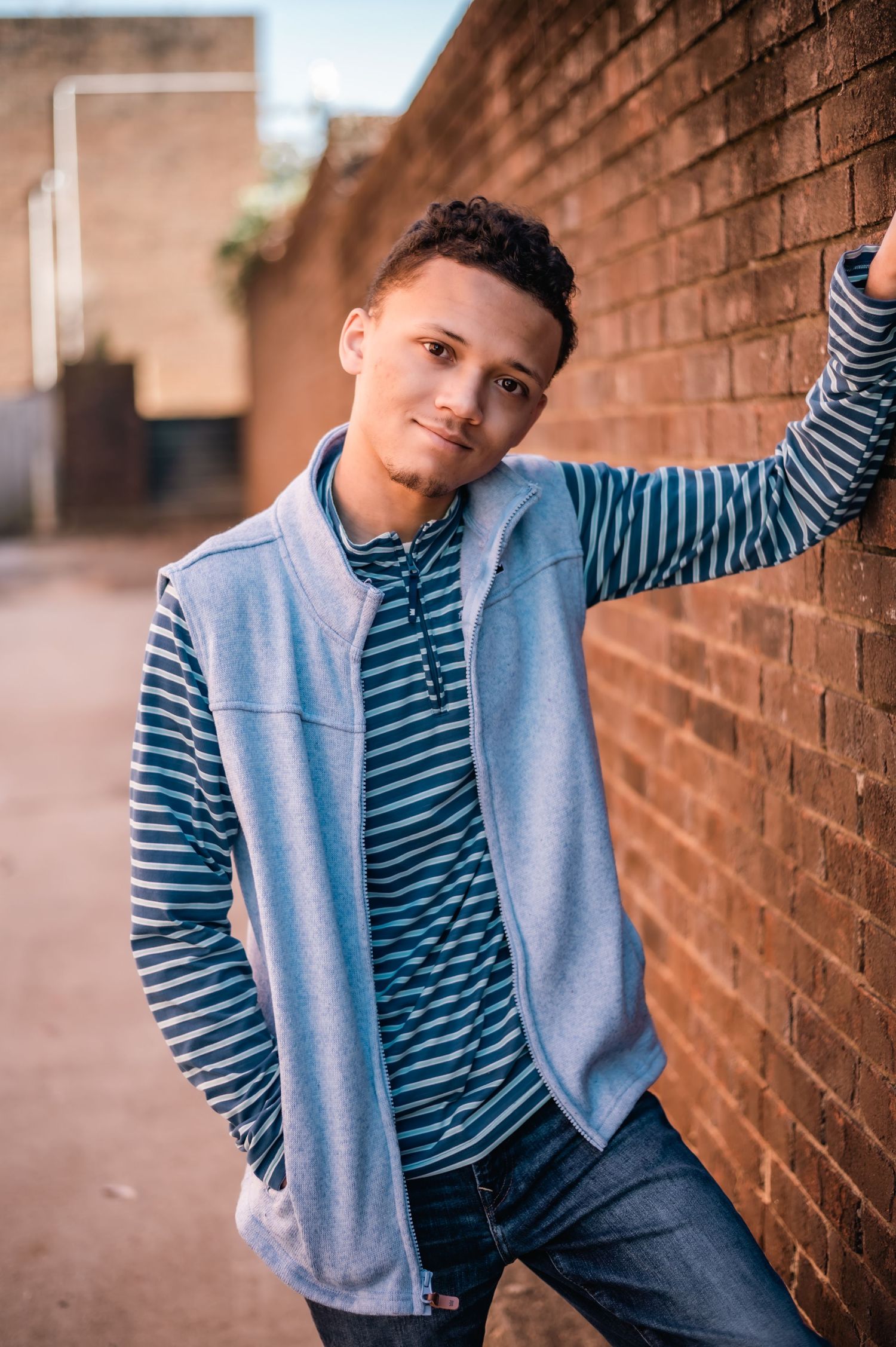 Young person in striped shirt and gray vest leaning against brick wall in casual outdoor portrait.
