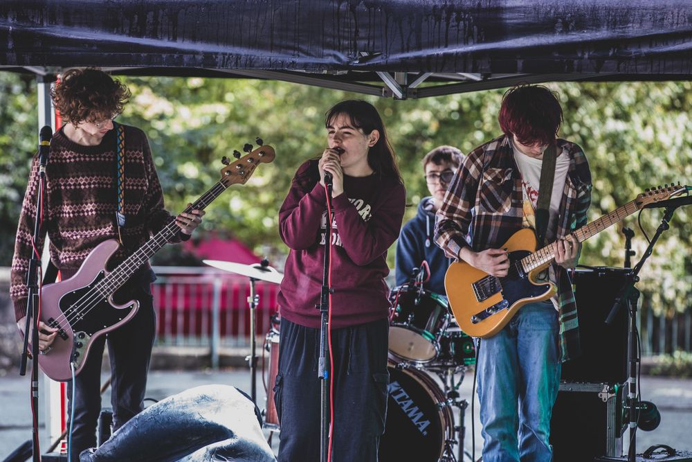 A rock band performs on an outdoor stage with guitarists and vocalist under a blue canopy.