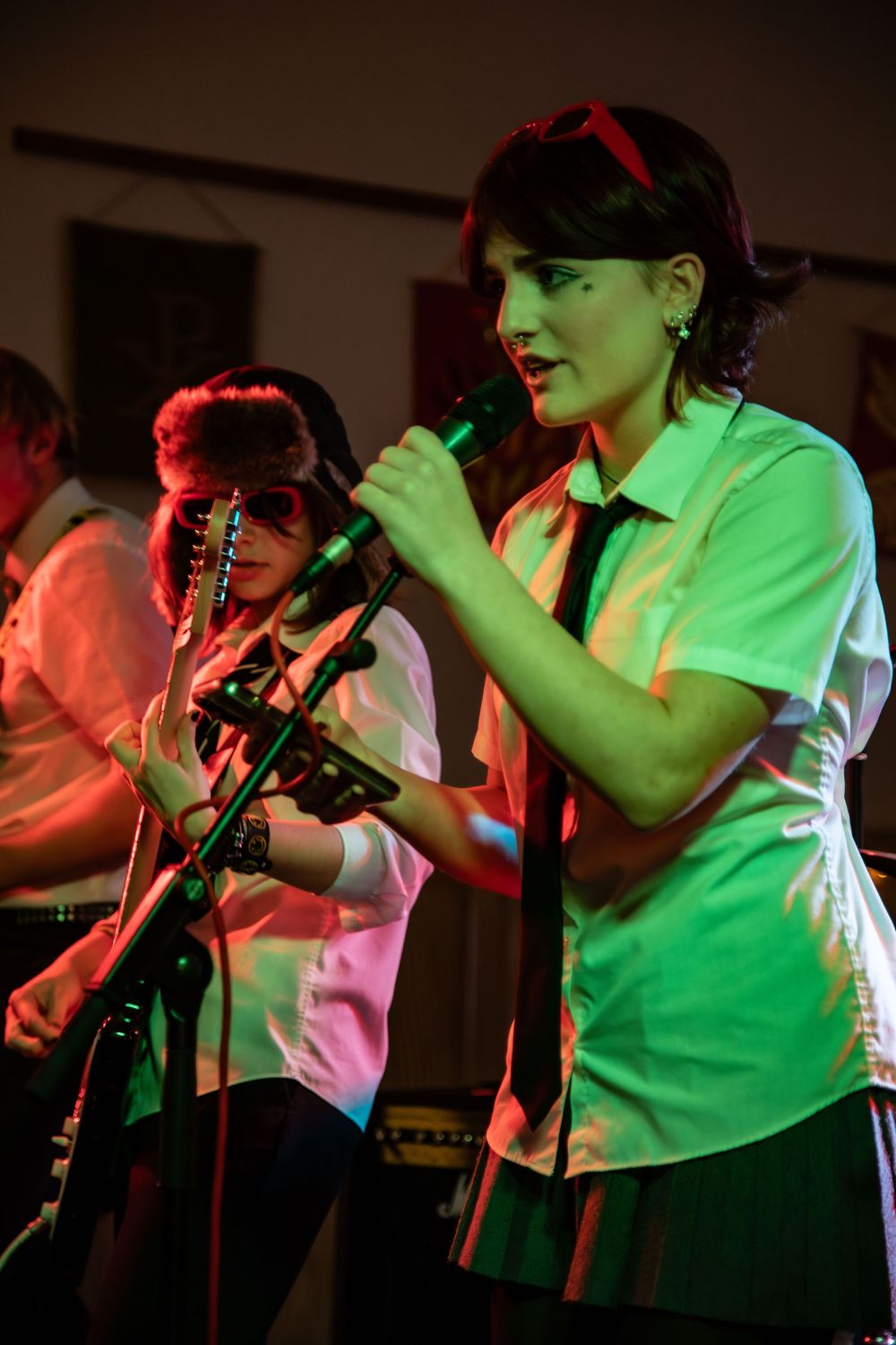 A performer in a green shirt and hat sings into a microphone on stage with colorful red and green lighting.