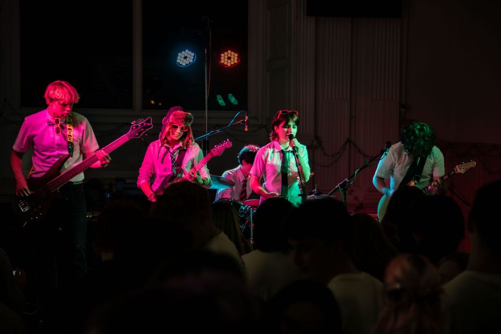 Band members perform on stage illuminated by vibrant pink and green lighting during a live concert performance.