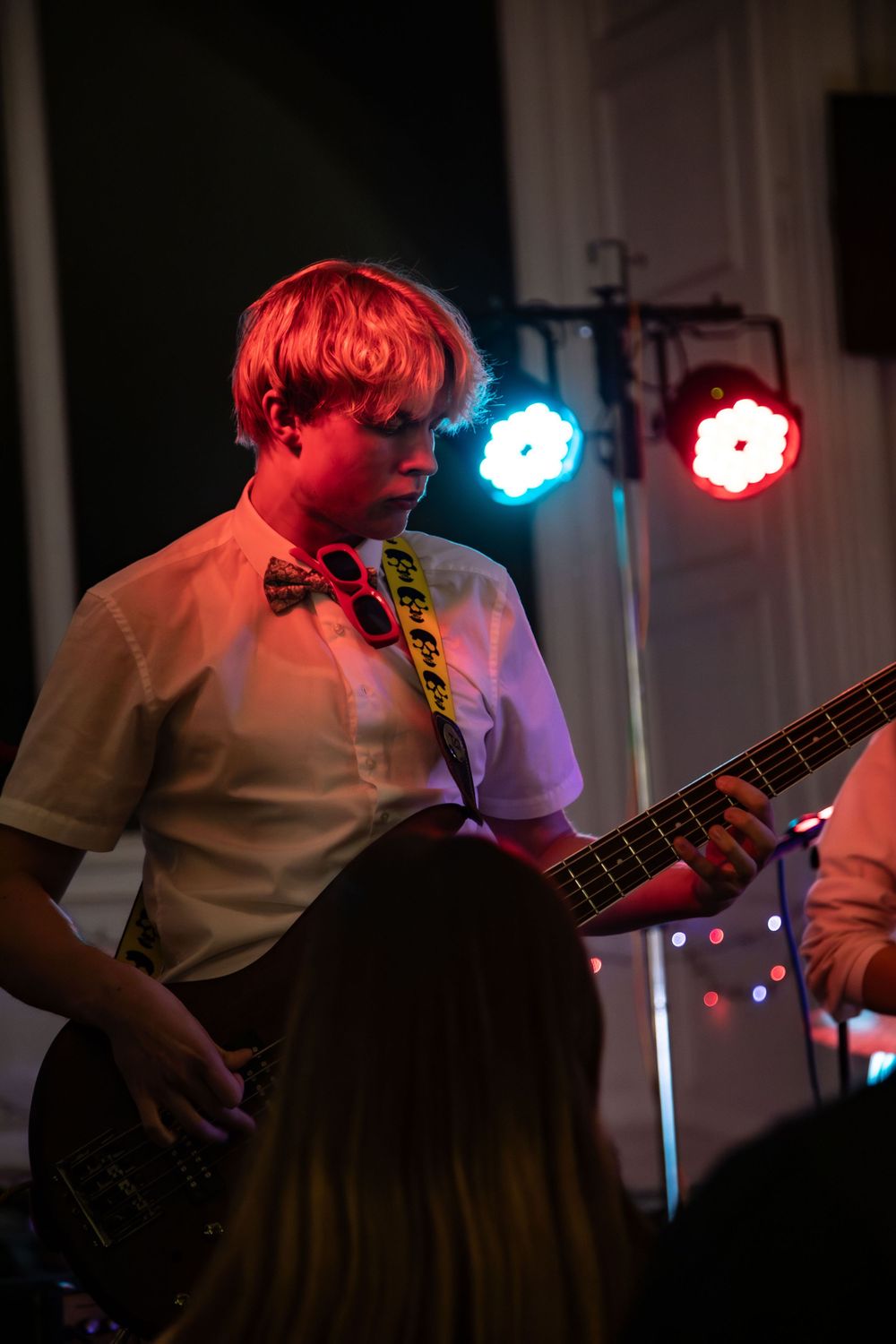 A musician in a white shirt and bow tie performs on stage with a bass guitar under red and blue stage lights.