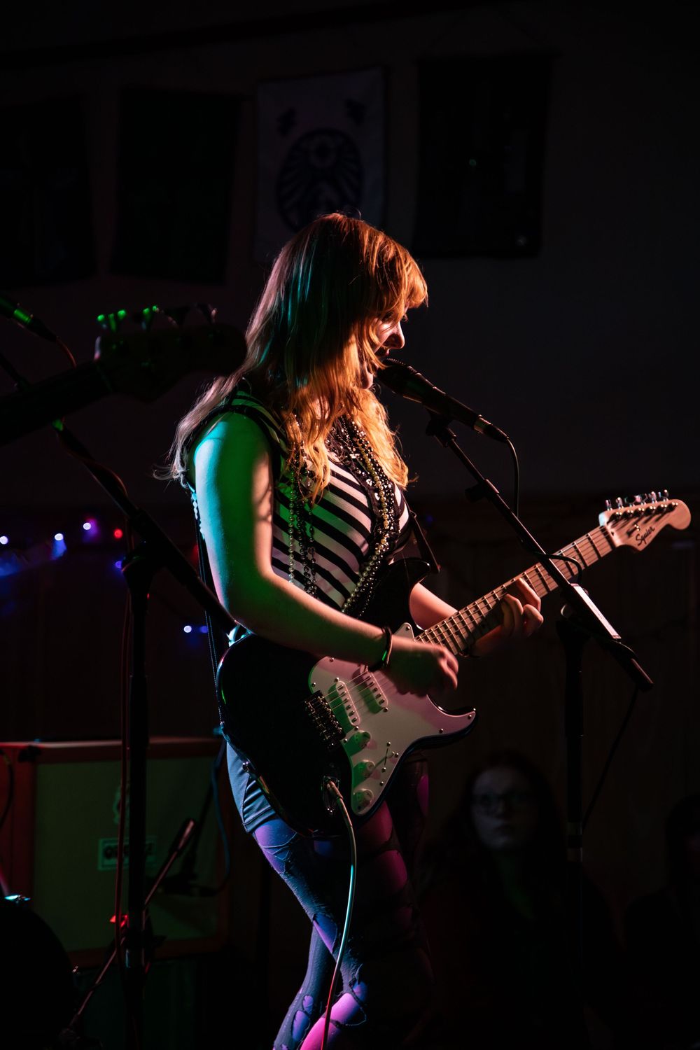 Female guitarist in striped top performs on stage with microphone and electric guitar under colorful stage lighting.