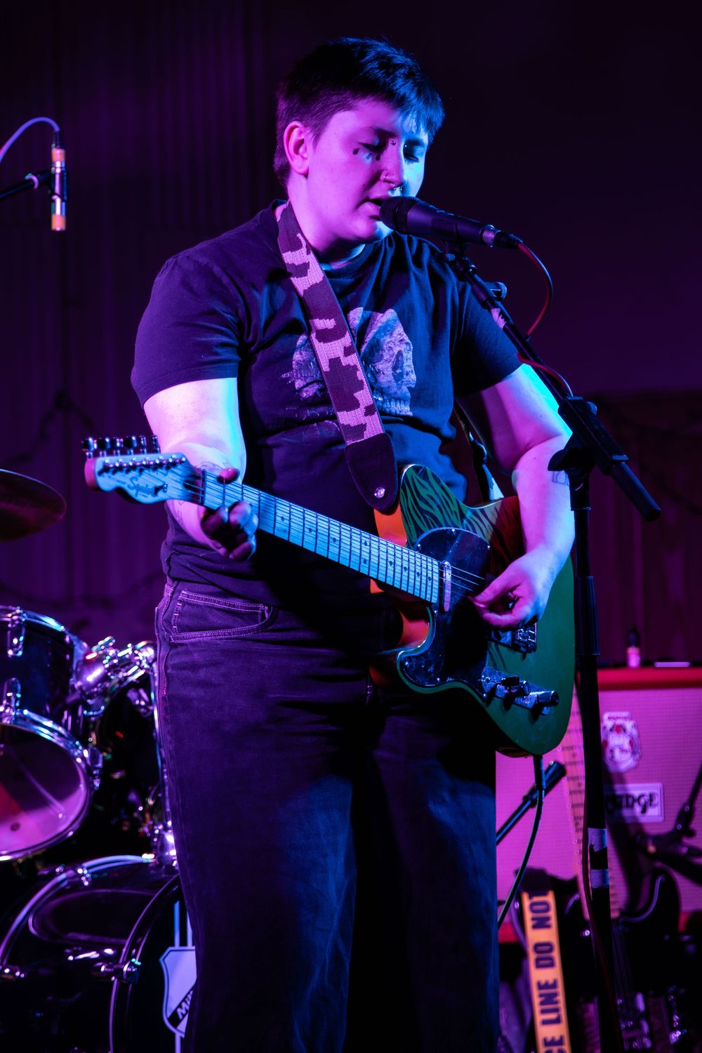 A musician performs on stage with a guitar, lit by blue lighting against a dark background, with a drum kit visible behind them.