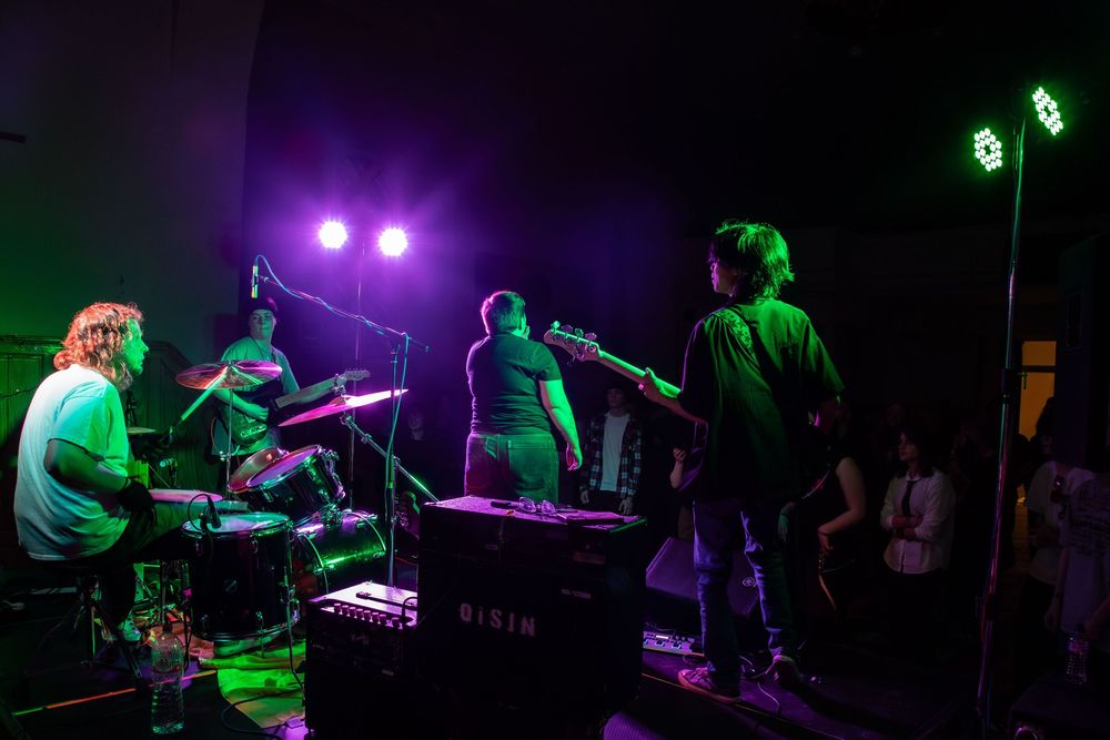 Musicians perform on a dimly lit stage with purple and green lighting during a live concert performance.