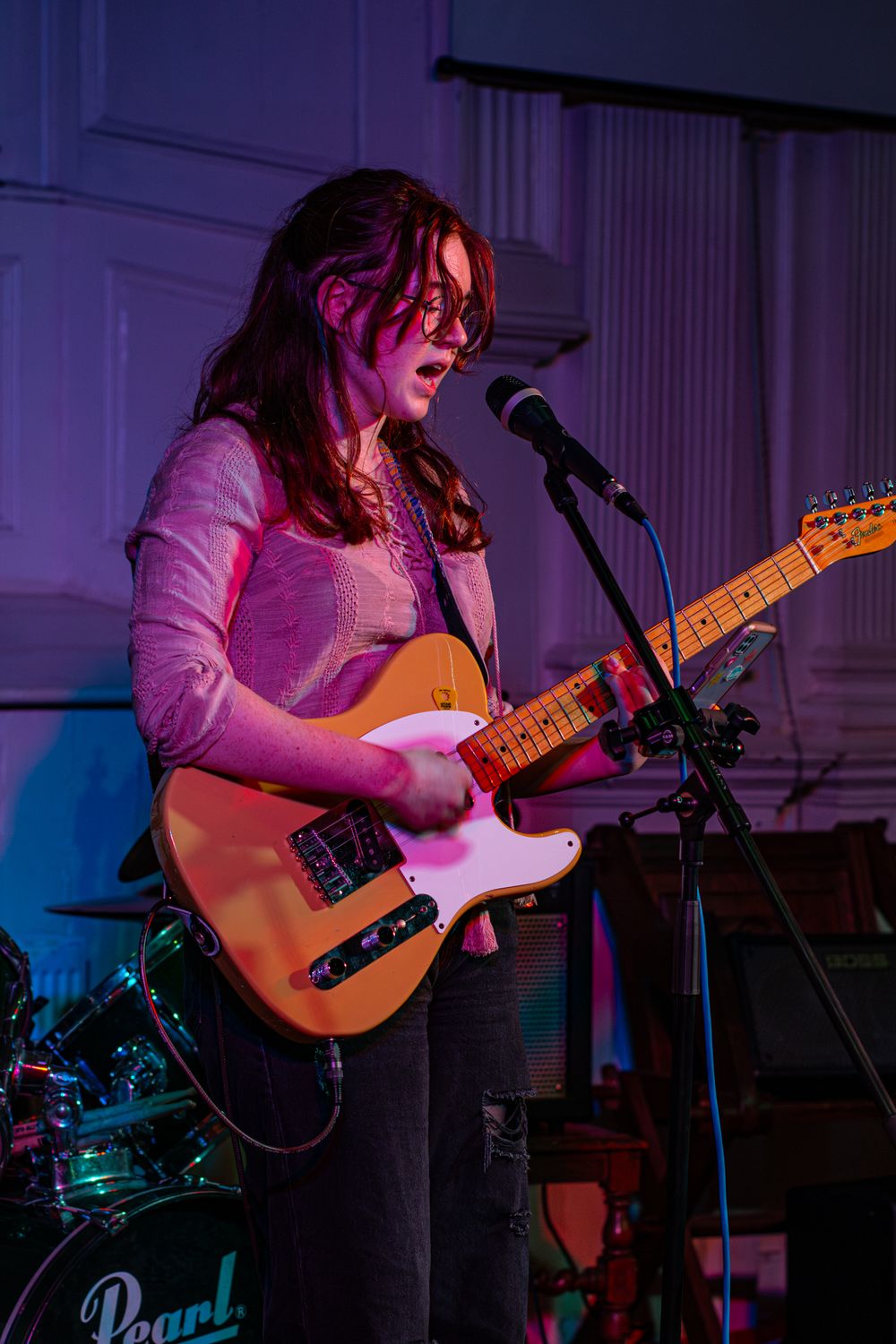 Musician performs on stage with a Telecaster guitar under purple and blue lighting while singing into a microphone.