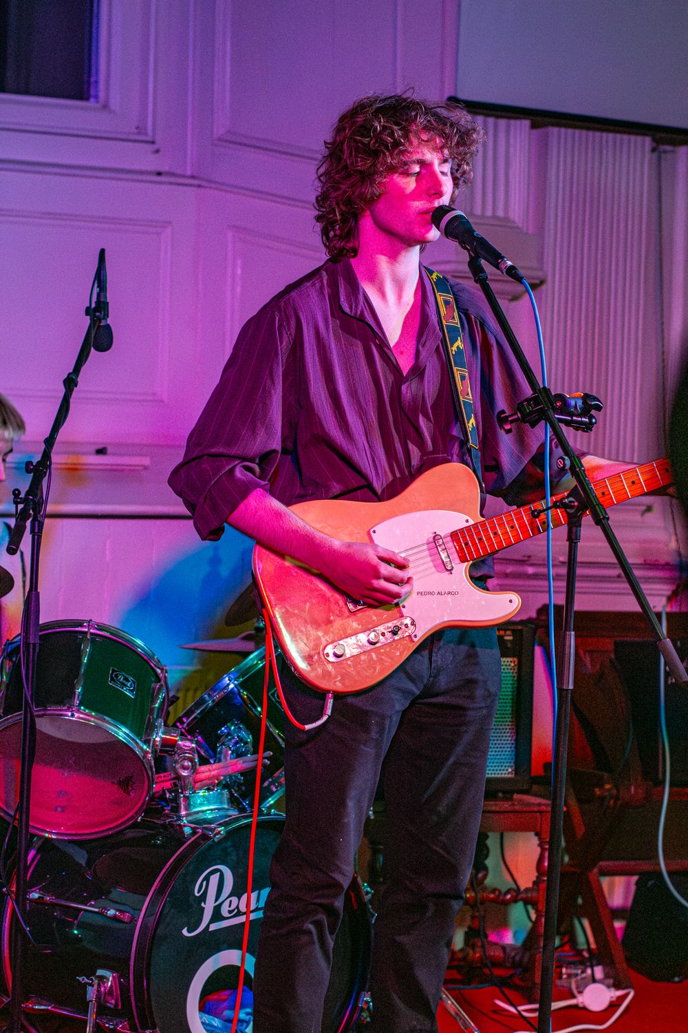 Musician performing on stage with a guitar under purple and blue concert lighting next to a Pearl drum kit.