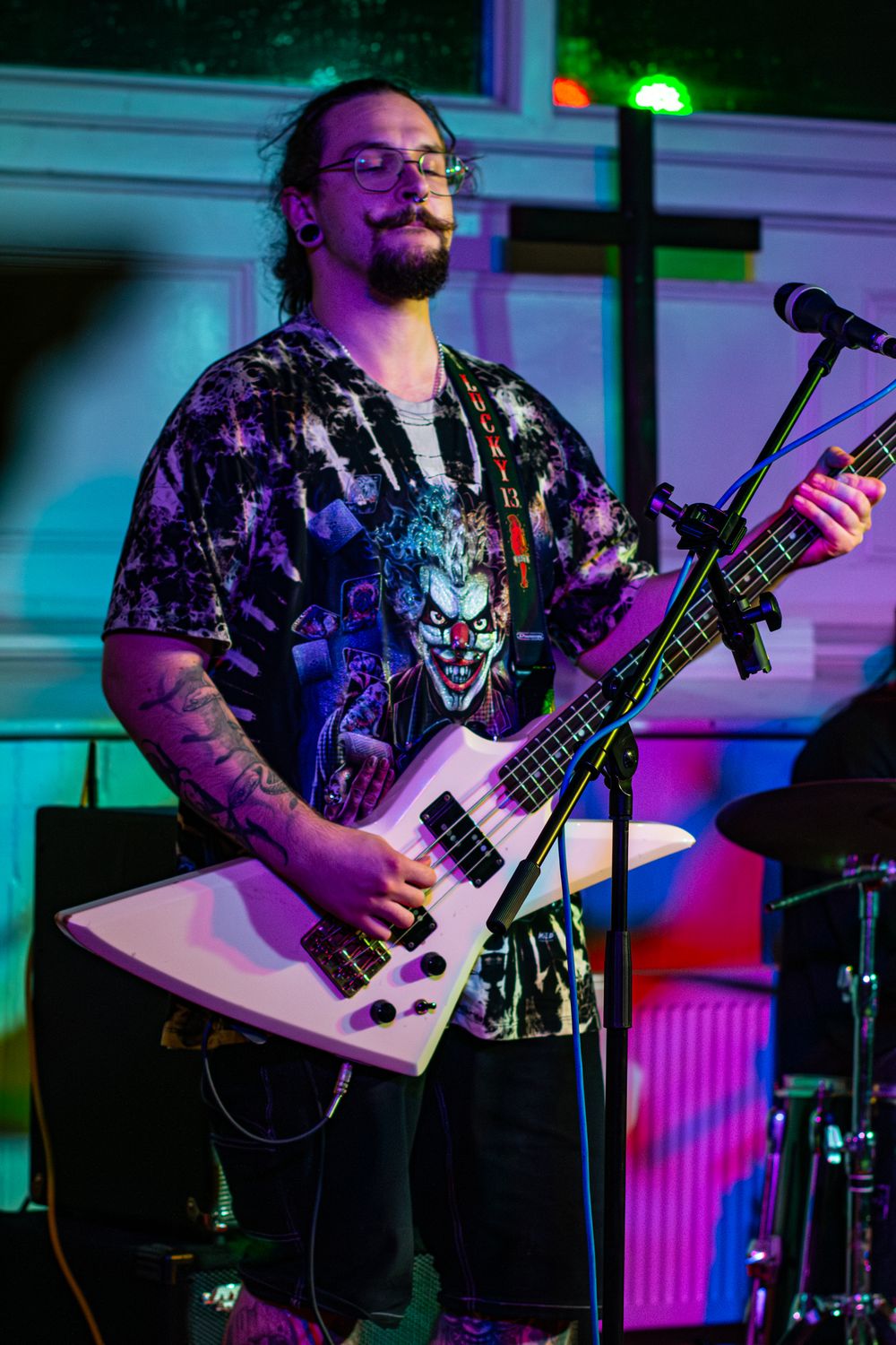 Musician in a Joker-themed shirt plays a white Explorer-style bass guitar on stage with colorful lighting.