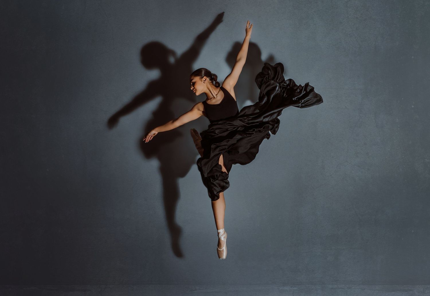 A dancer in a black leotard performs a graceful pose with one leg extended upward against a dark backdrop.