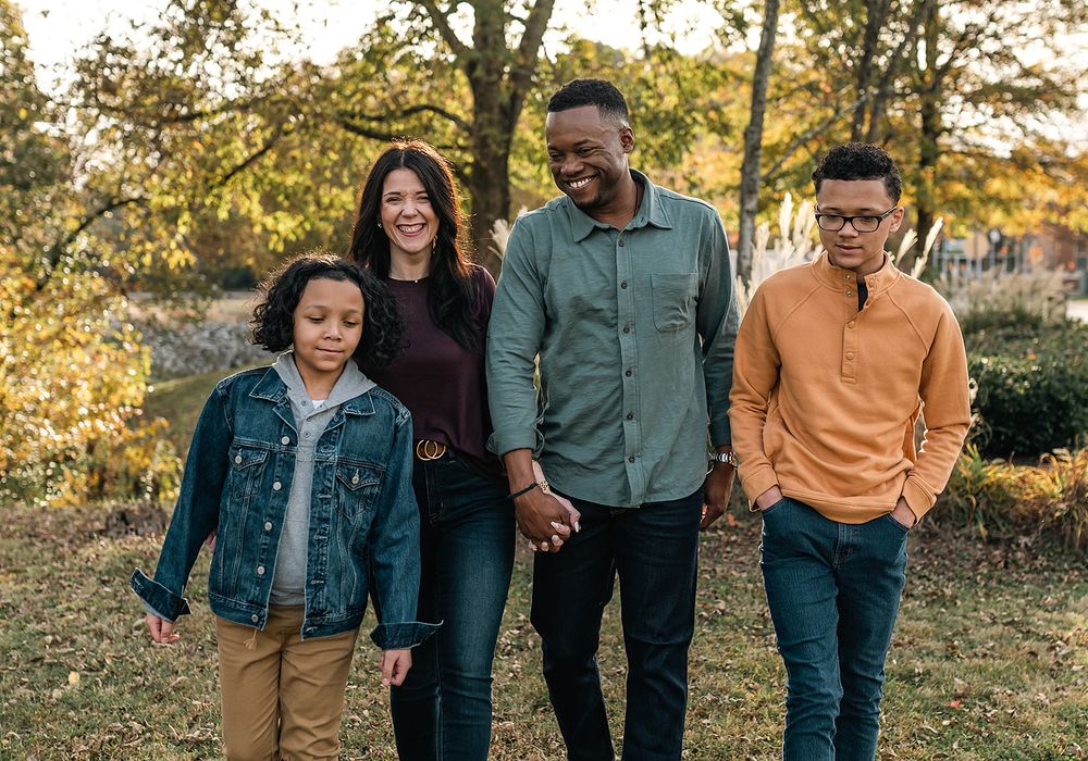 A family walks together outdoors during golden hour with autumn trees in the background.