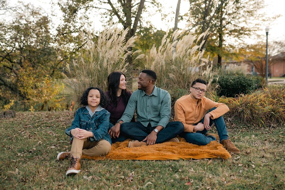A family sits together on an orange blanket surrounded by autumn foliage and ornamental grasses.