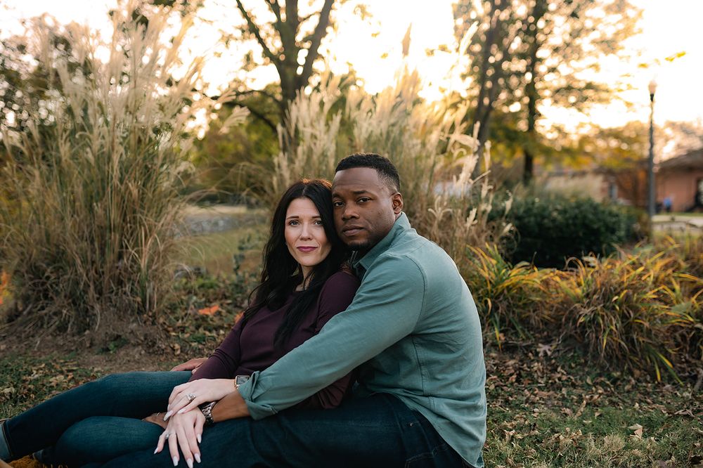A couple sits together outdoors at sunset with ornamental grasses and trees in the background.