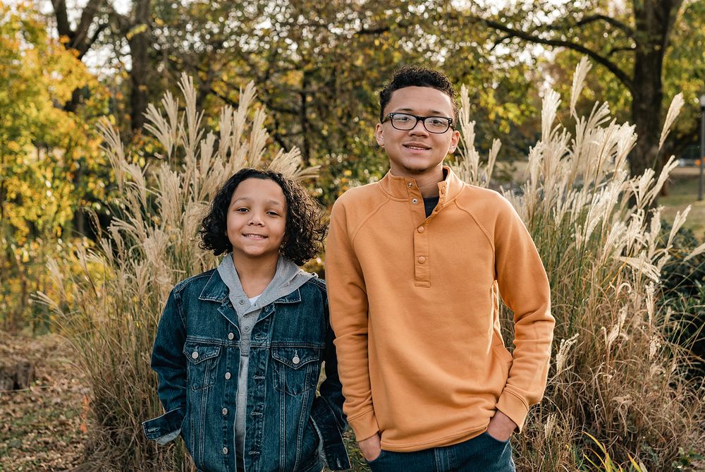 Two people stand together outdoors with autumn foliage and decorative grasses in the background.