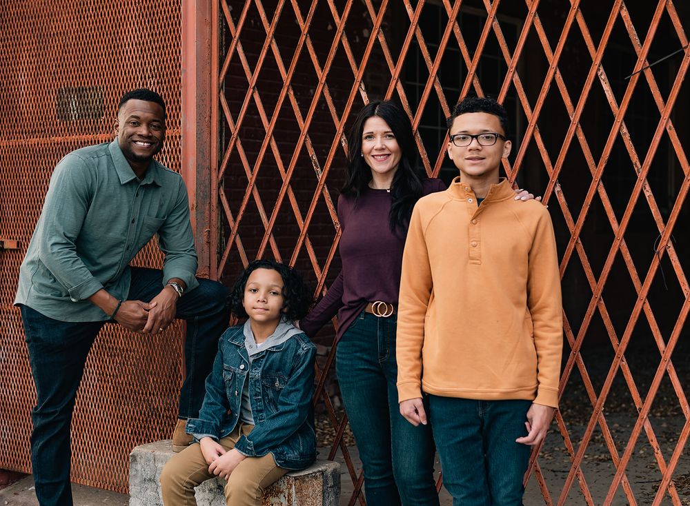 A family poses in front of a red brick wall with diamond pattern metal grating.