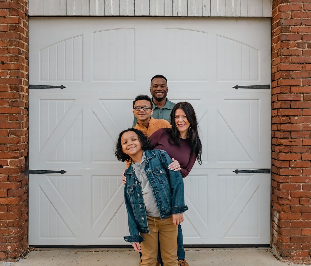 A family poses playfully in front of a white garage door with brick columns.