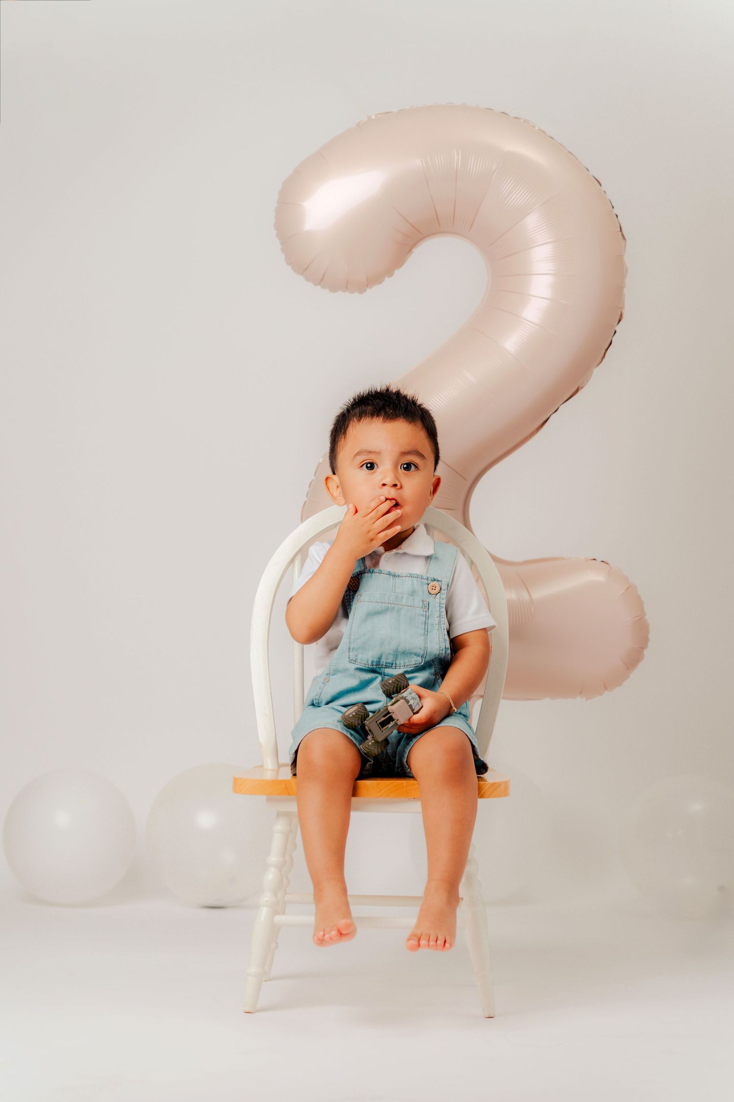 Three photos of a small child in blue clothing sitting on a white chair with a large number 2 balloon decoration.
