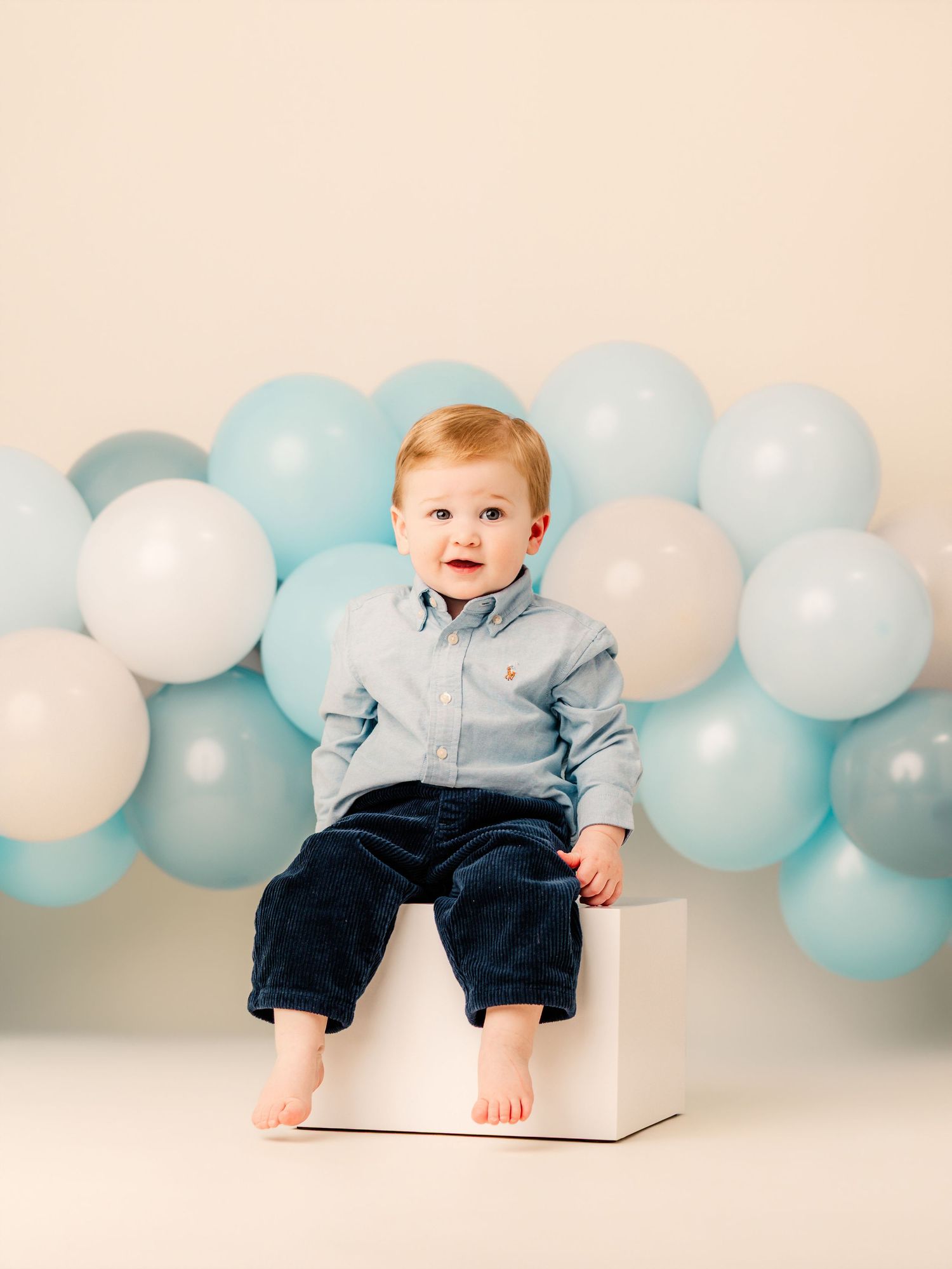 A baby boy in a blue shirt and navy pants sits on a white cube surrounded by pastel blue and white balloons.