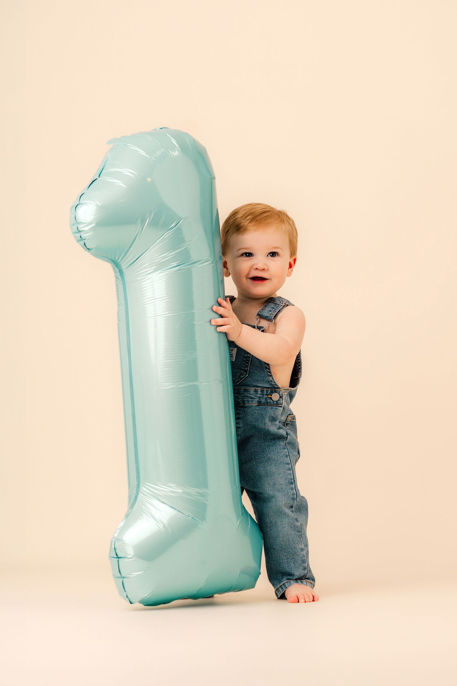 Smiling toddler in denim overalls holding a large teal number 1 balloon on a beige background.