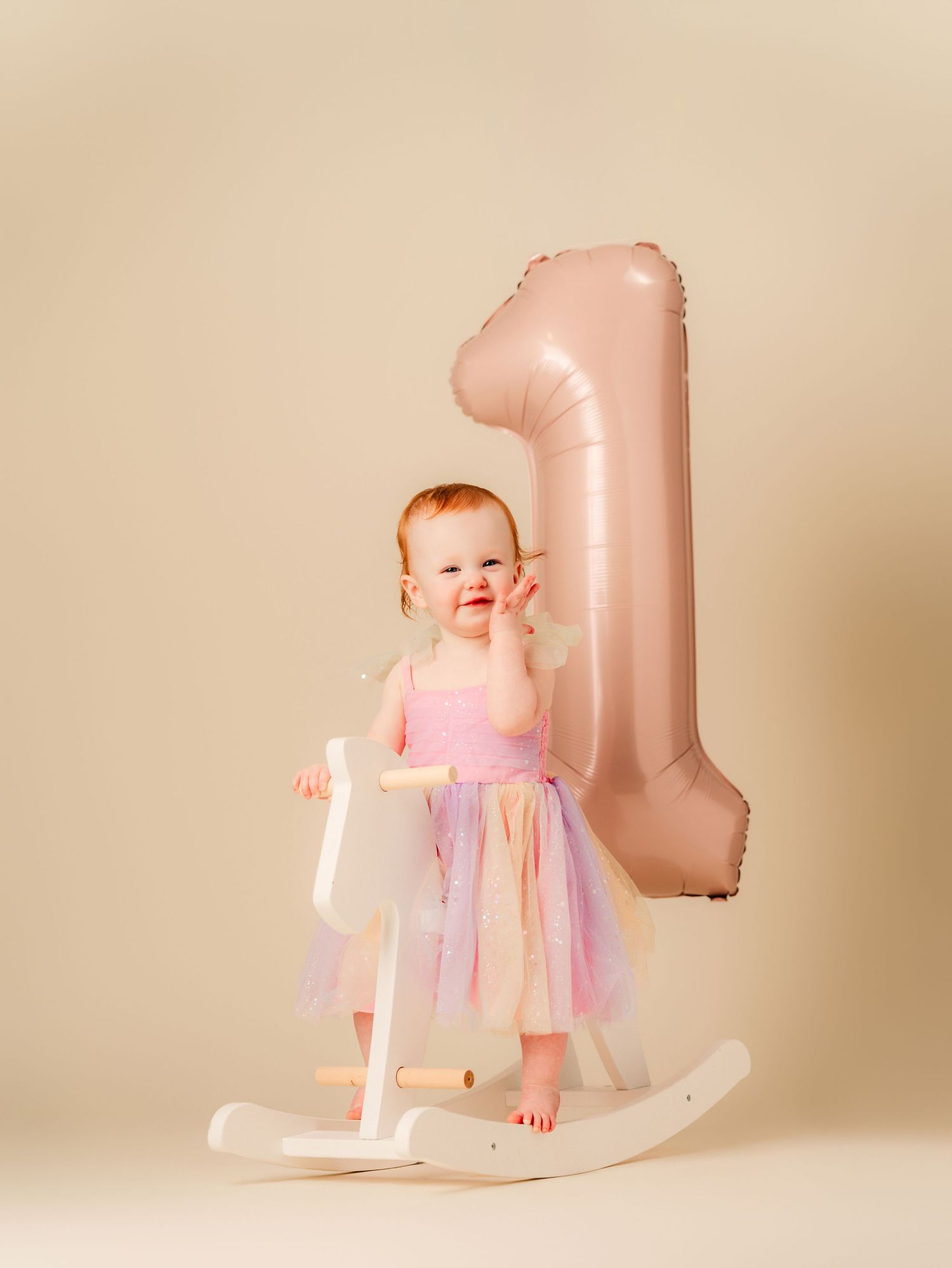 A smiling baby girl in a pink tutu dress stands by a rocking horse with a large number 1 balloon for her first birthday.