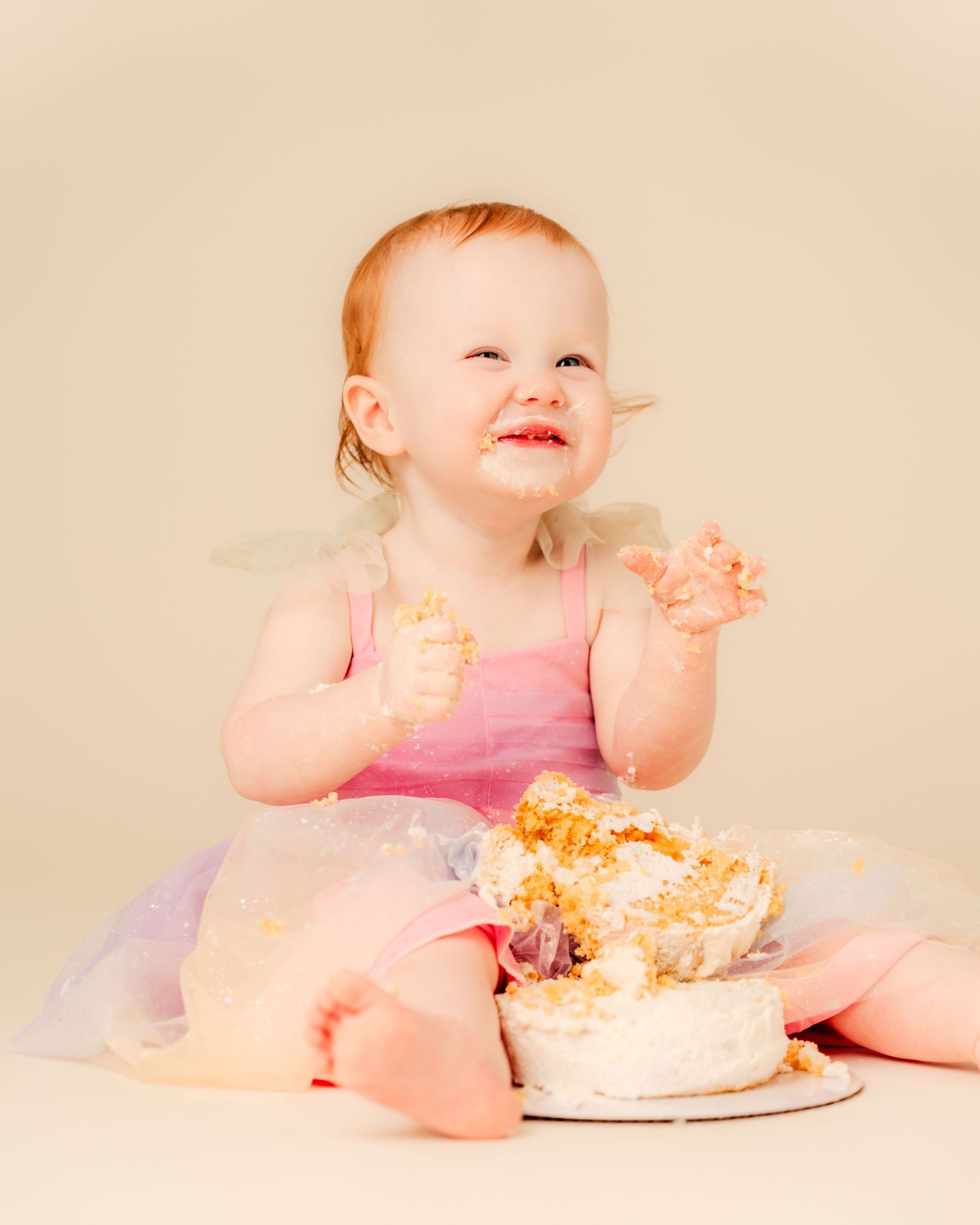 Baby girl in pink tutu smashes first birthday cake, laughing with frosting on her face and hands.