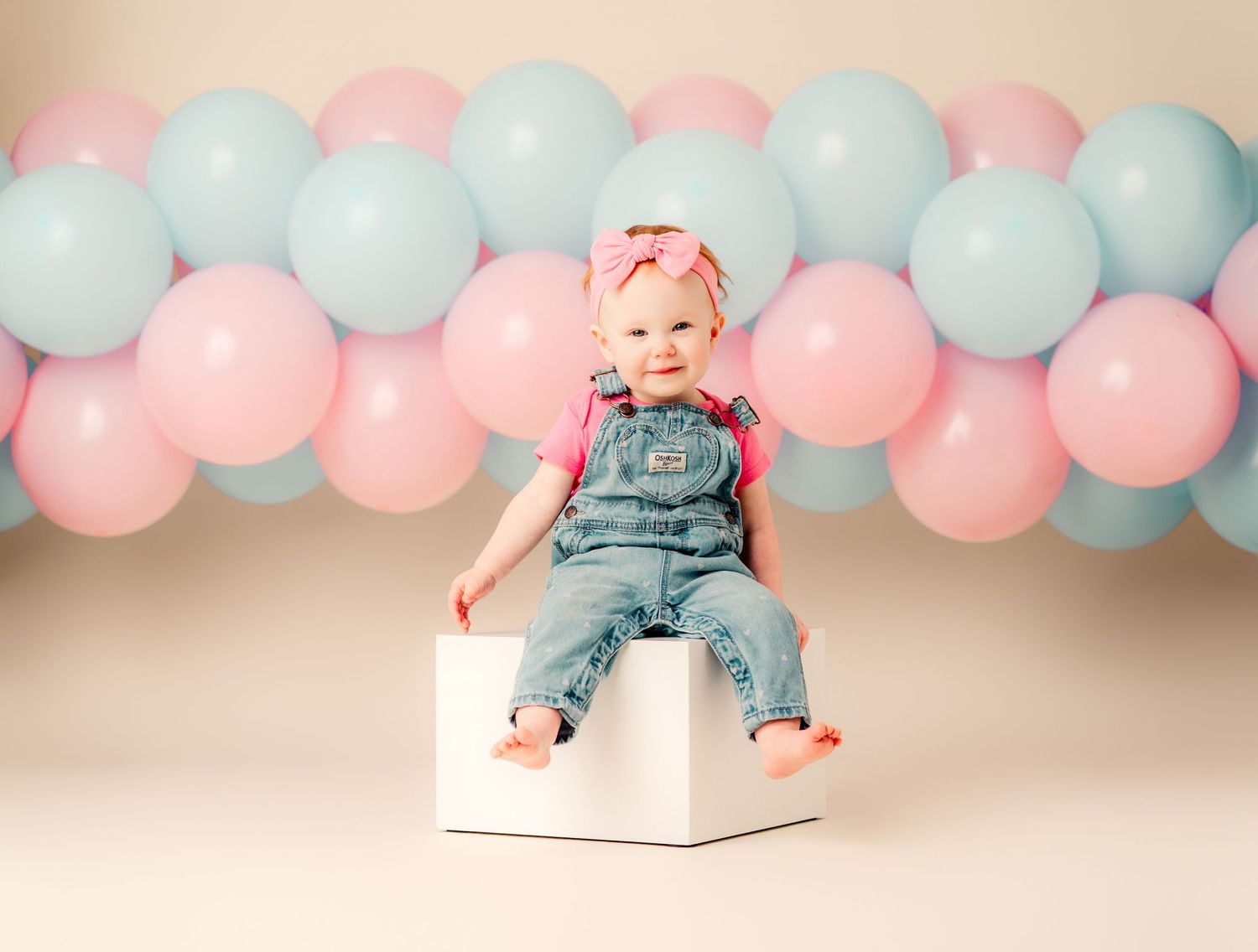 Baby girl in denim overalls sits on white cube surrounded by pink and blue balloons for a gender reveal photo.