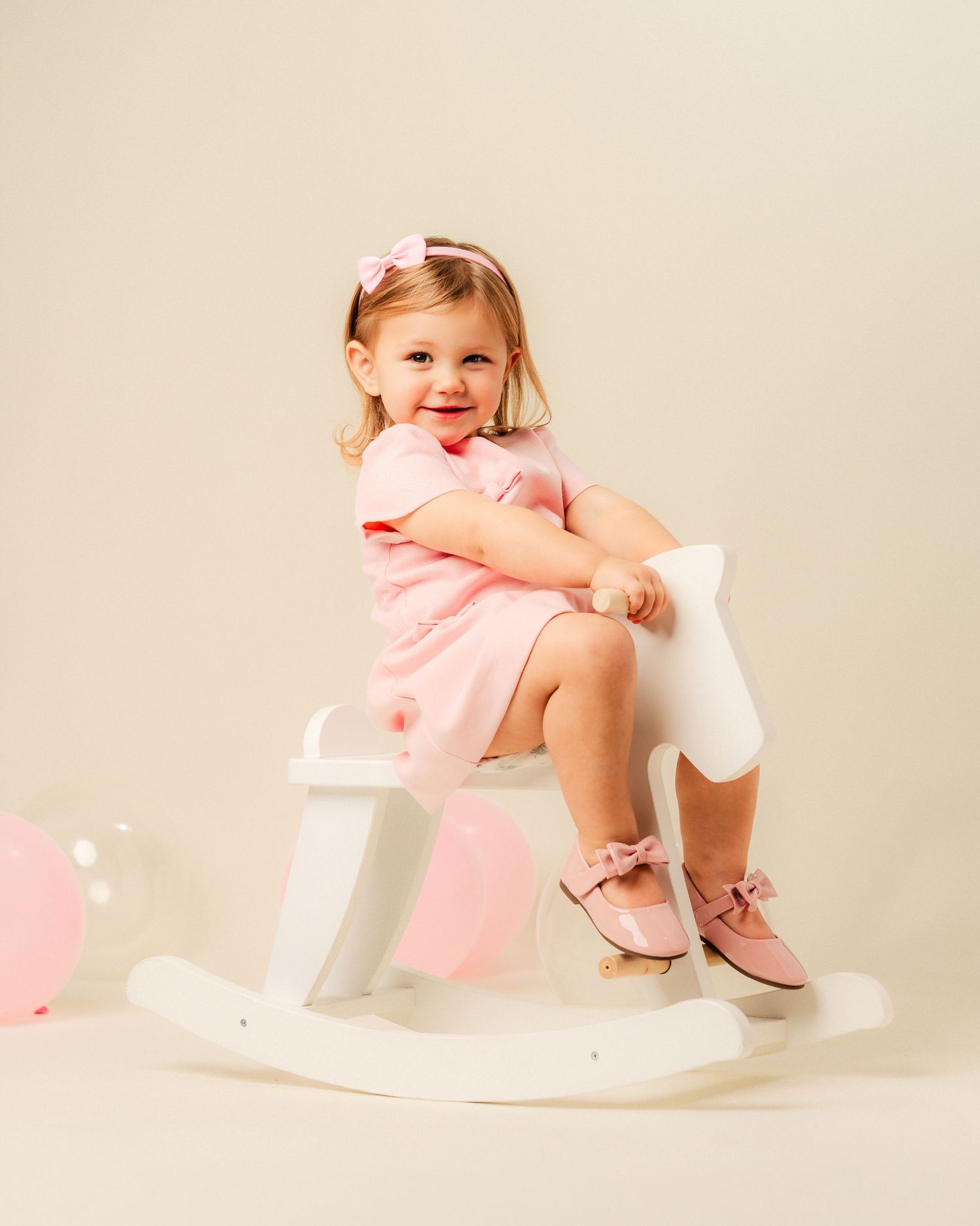 A small child in a pink dress sits on a white rocking horse against a neutral background with pink balloons.
