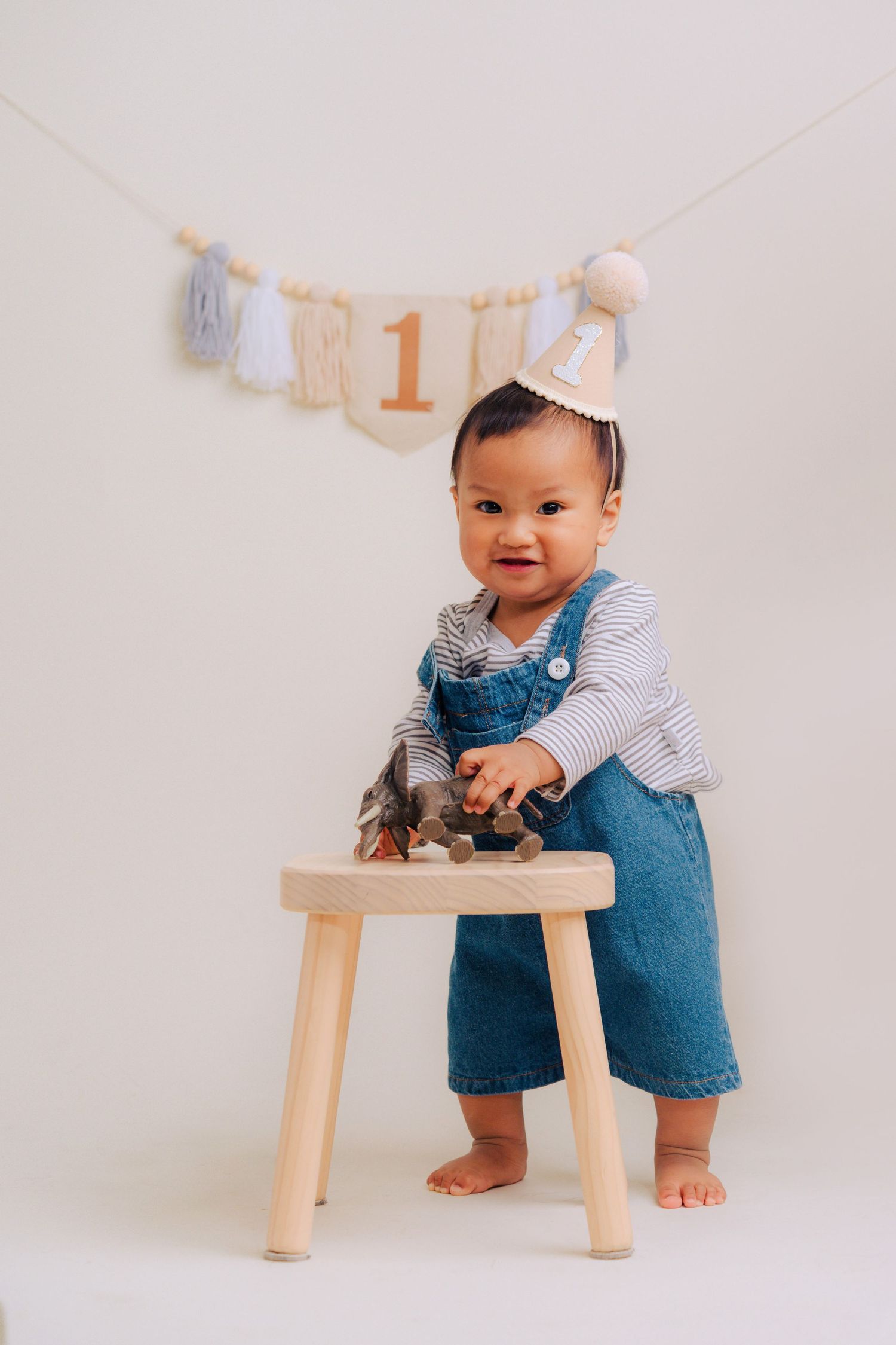 A small child in denim overalls and striped shirt stands by a wooden stool with first birthday decorations hung above.