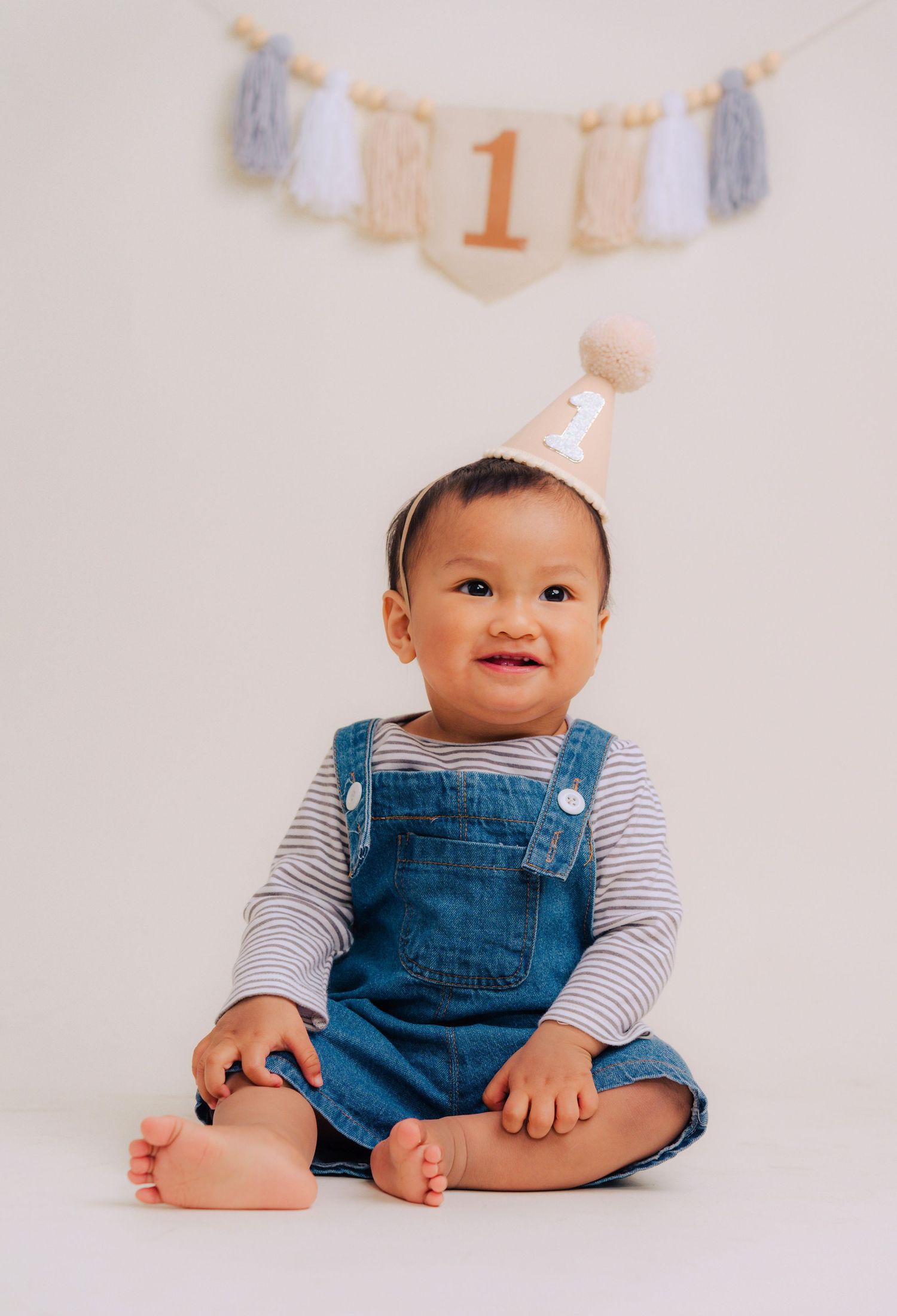 Small child wearing denim overalls and striped shirt sits on white backdrop with first birthday banner decoration above.