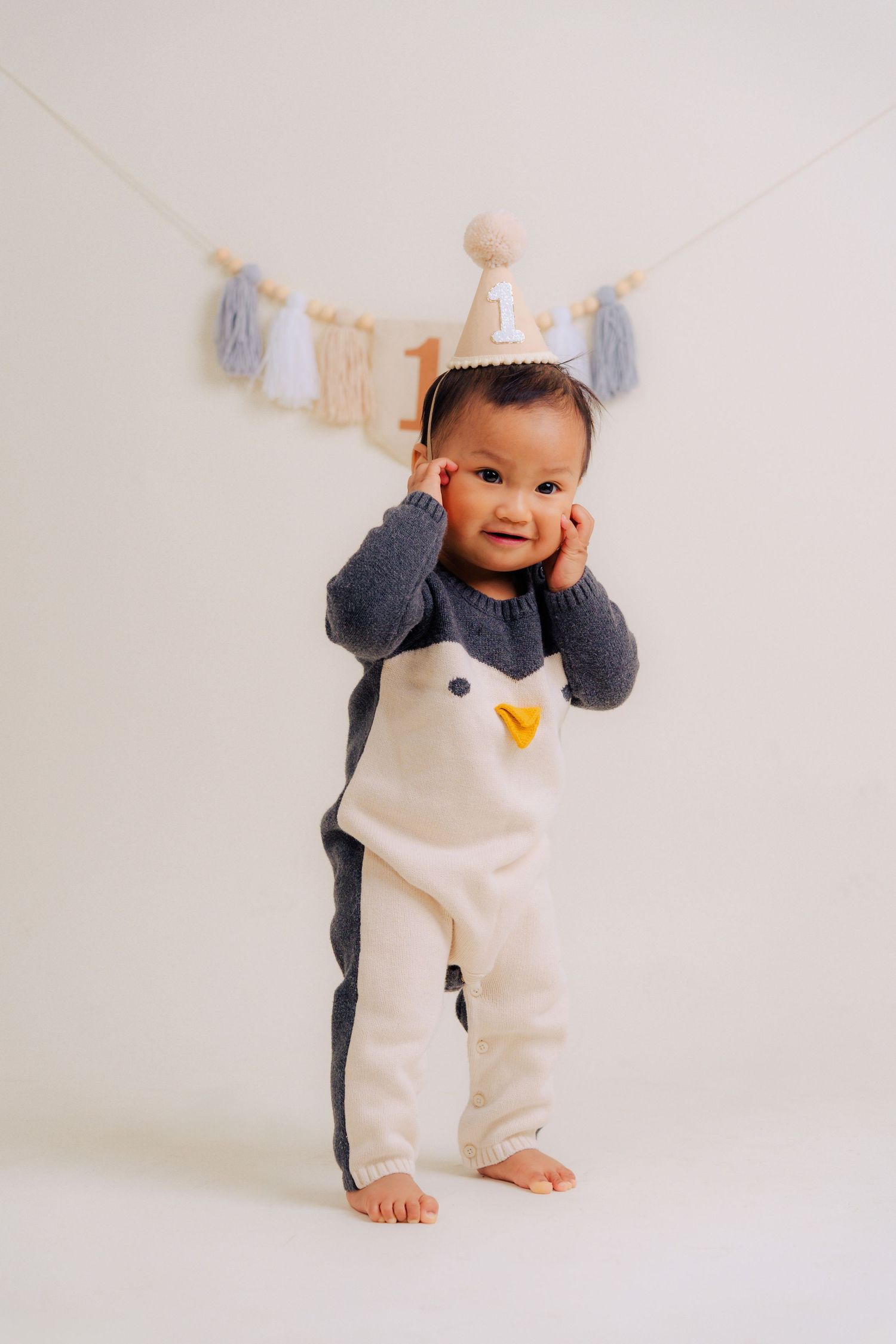 A small child wearing a black and white penguin-themed romper stands barefoot against a white background with baby clothes hanging above.