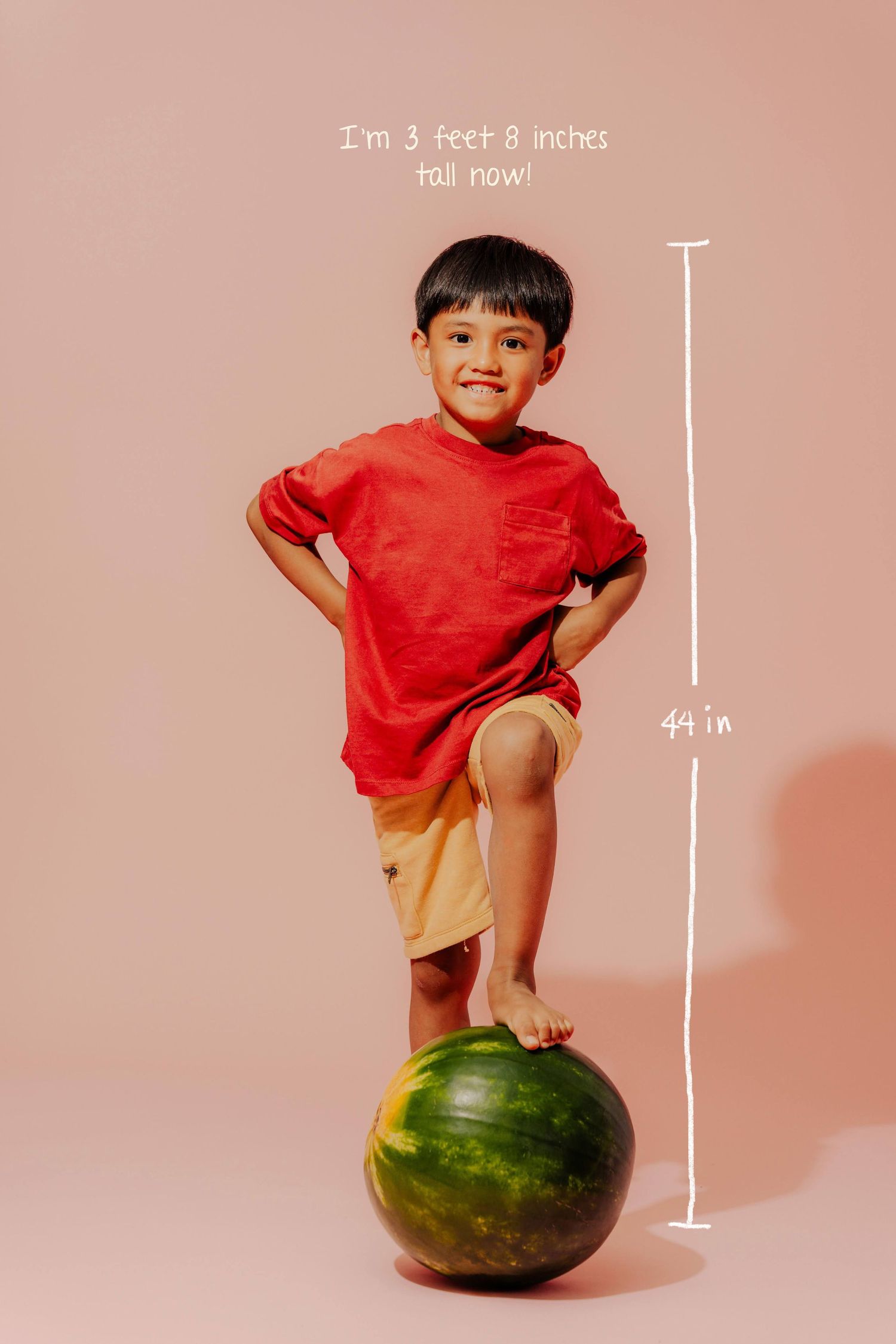 A playful child balancing on a large watermelon against a pink background with height measurements shown.
