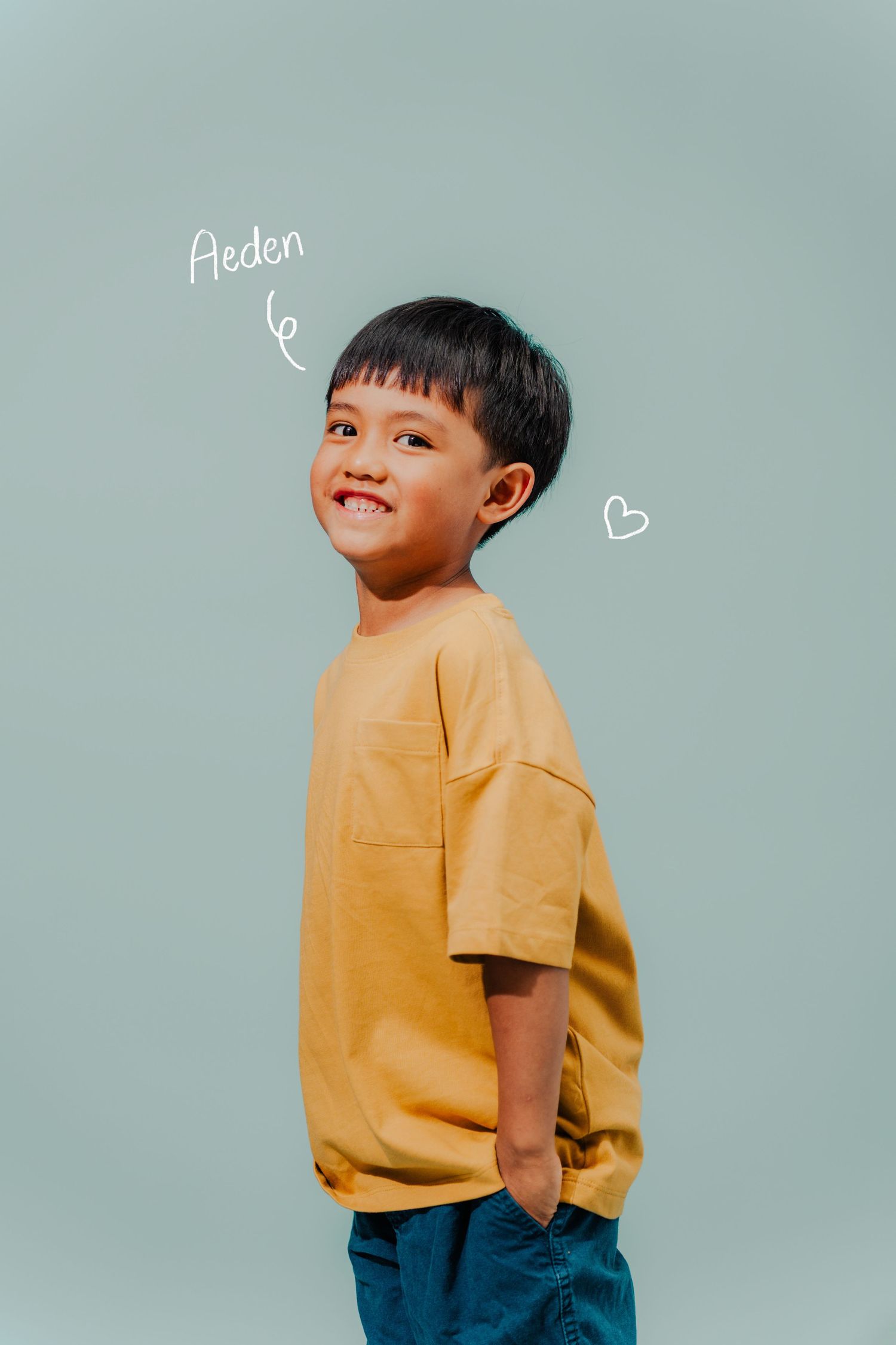 Portrait of a child in yellow t-shirt smiling against mint green background with handwritten doodles.