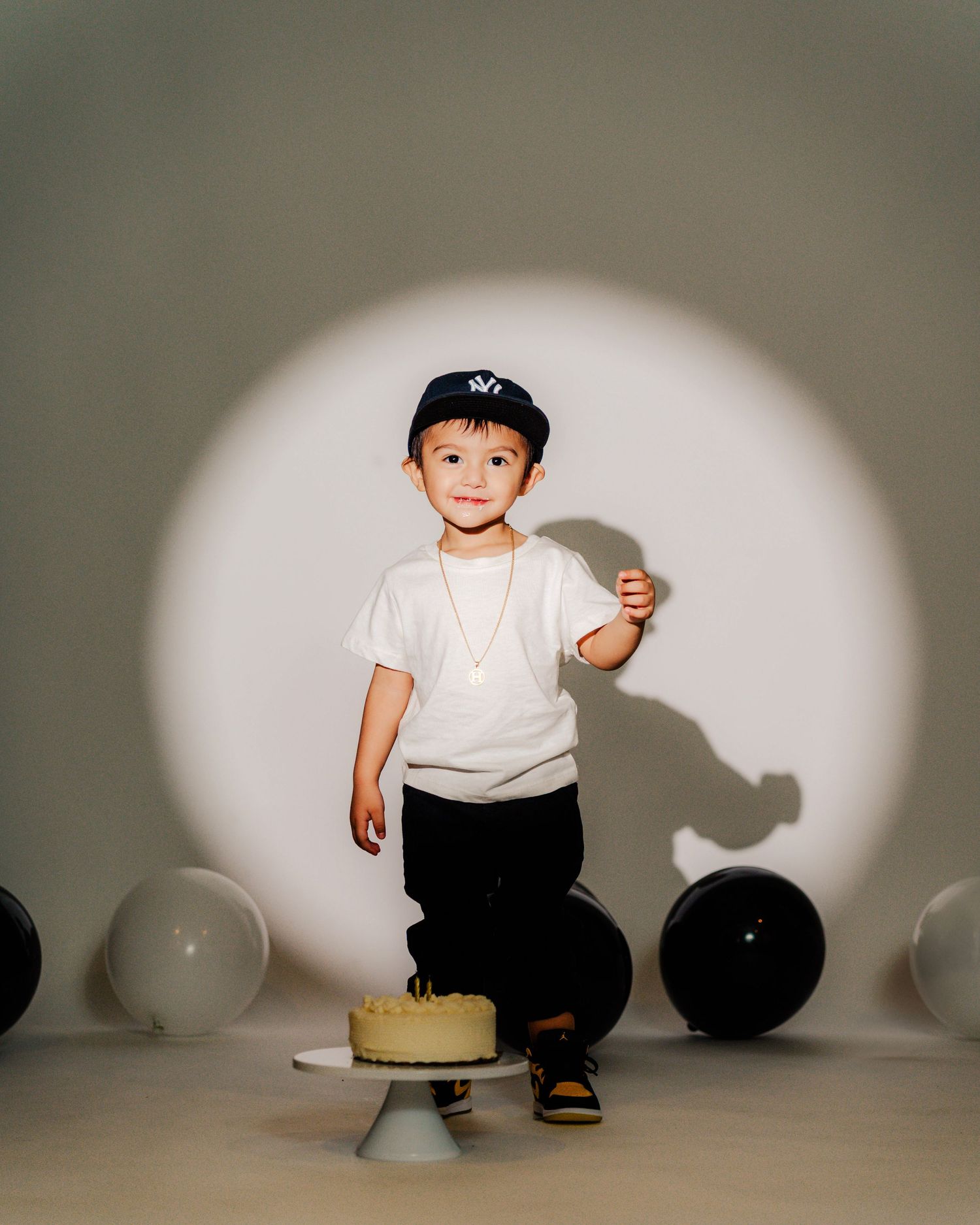 Small child crouches down near white balloons against a gray wall in an artistic minimalist scene.