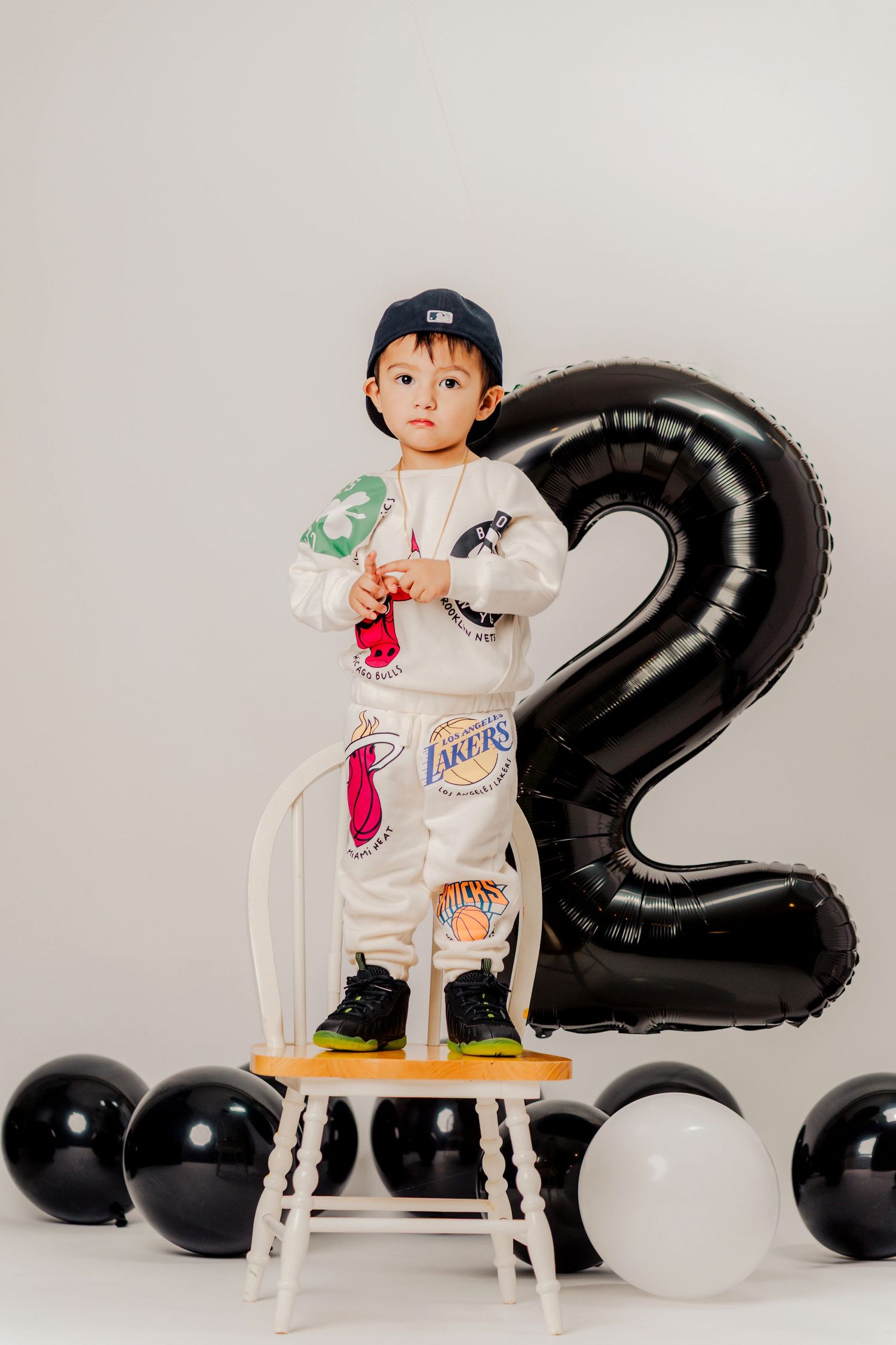 Child sitting on white chair surrounded by black and white balloons with number 2 decoration for birthday celebration.
