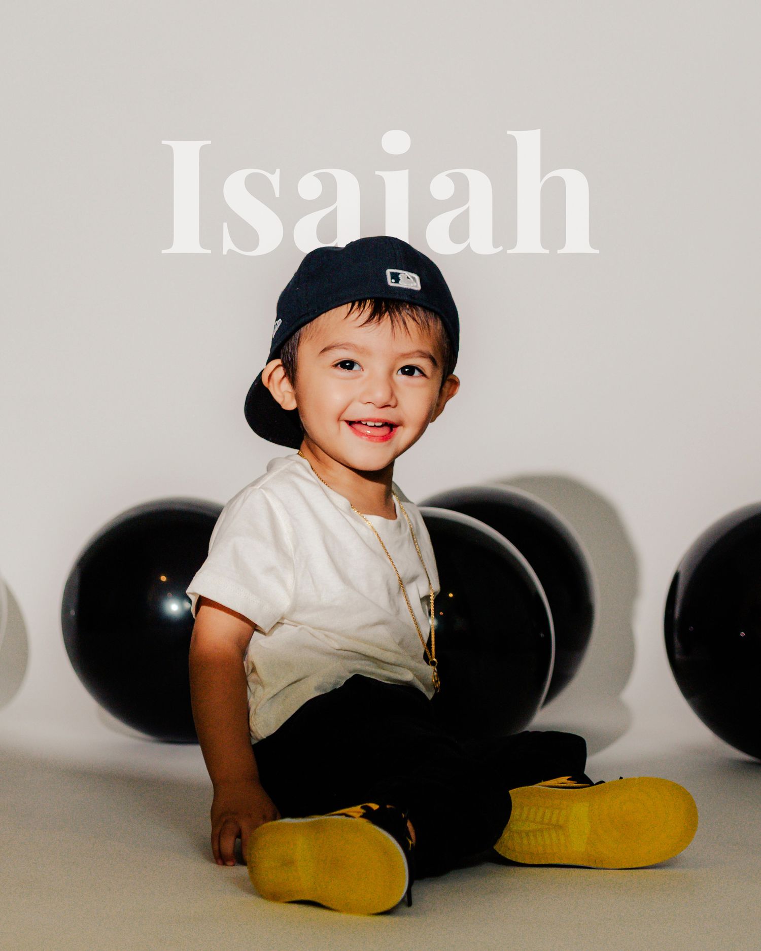 A child poses against a white backdrop with black balloons and yellow boots in a studio photography session.