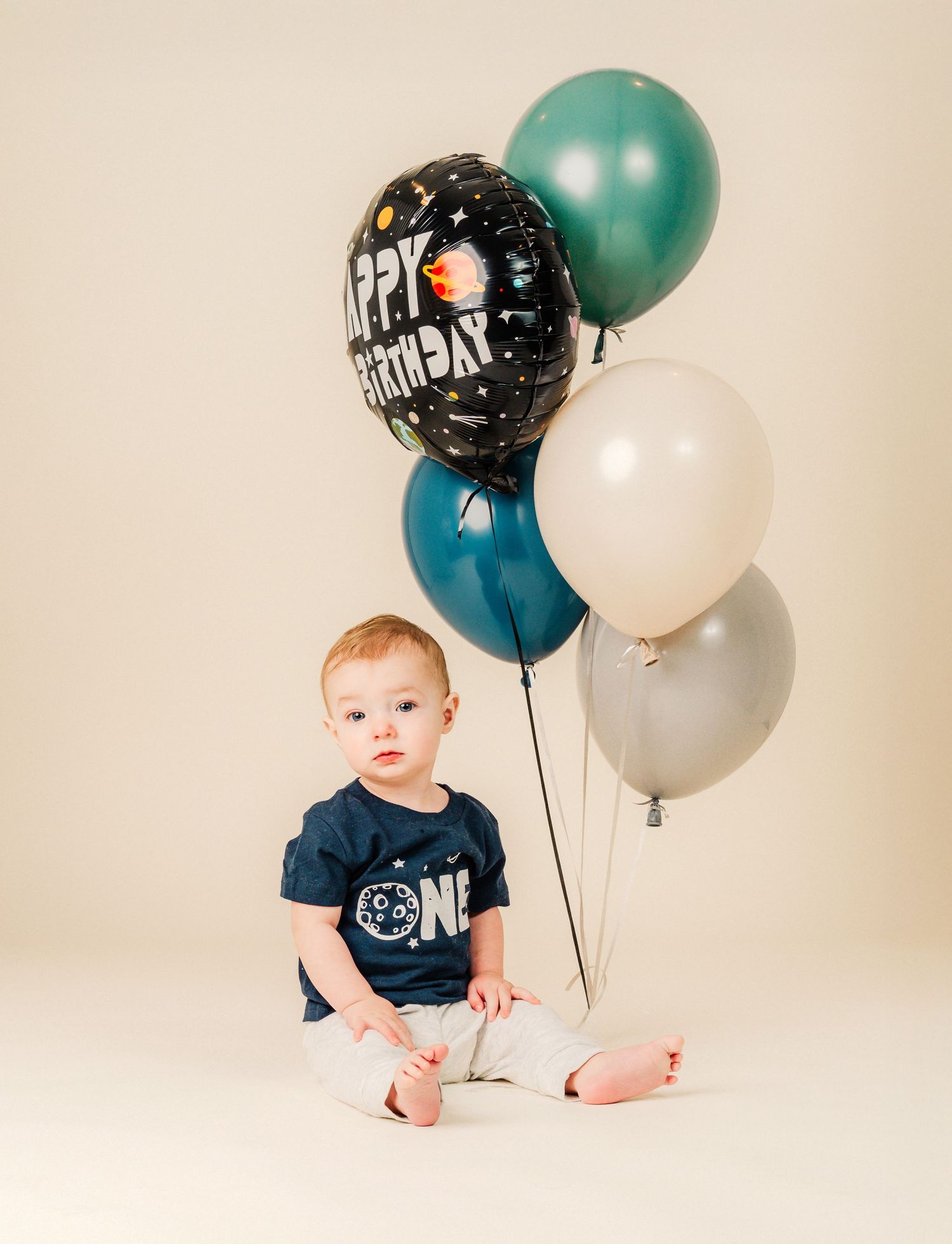 A small child in a blue shirt sits among green, white and black birthday balloons against a neutral background.