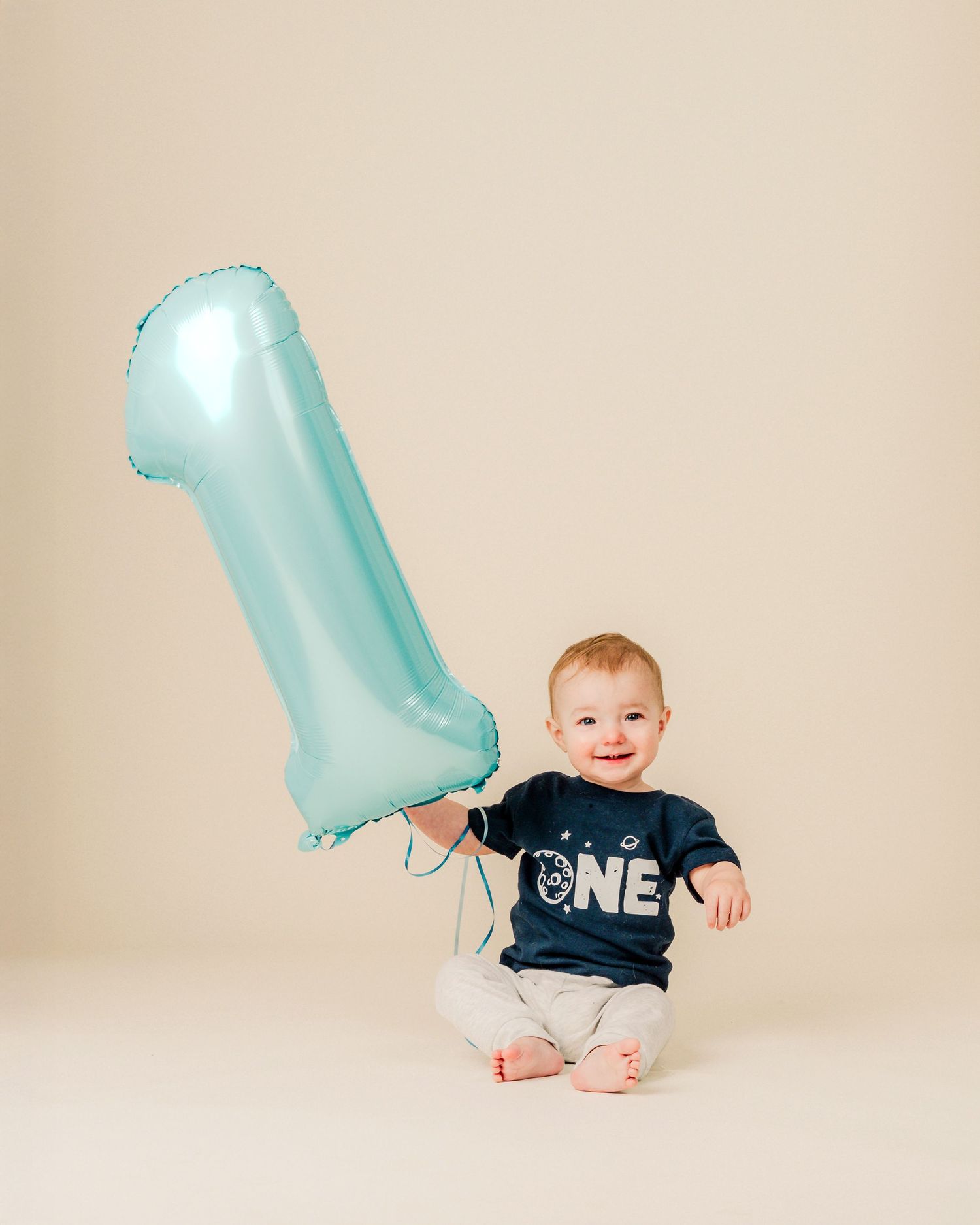 Baby in navy shirt sits on cream backdrop with large blue number one balloon for first birthday celebration.