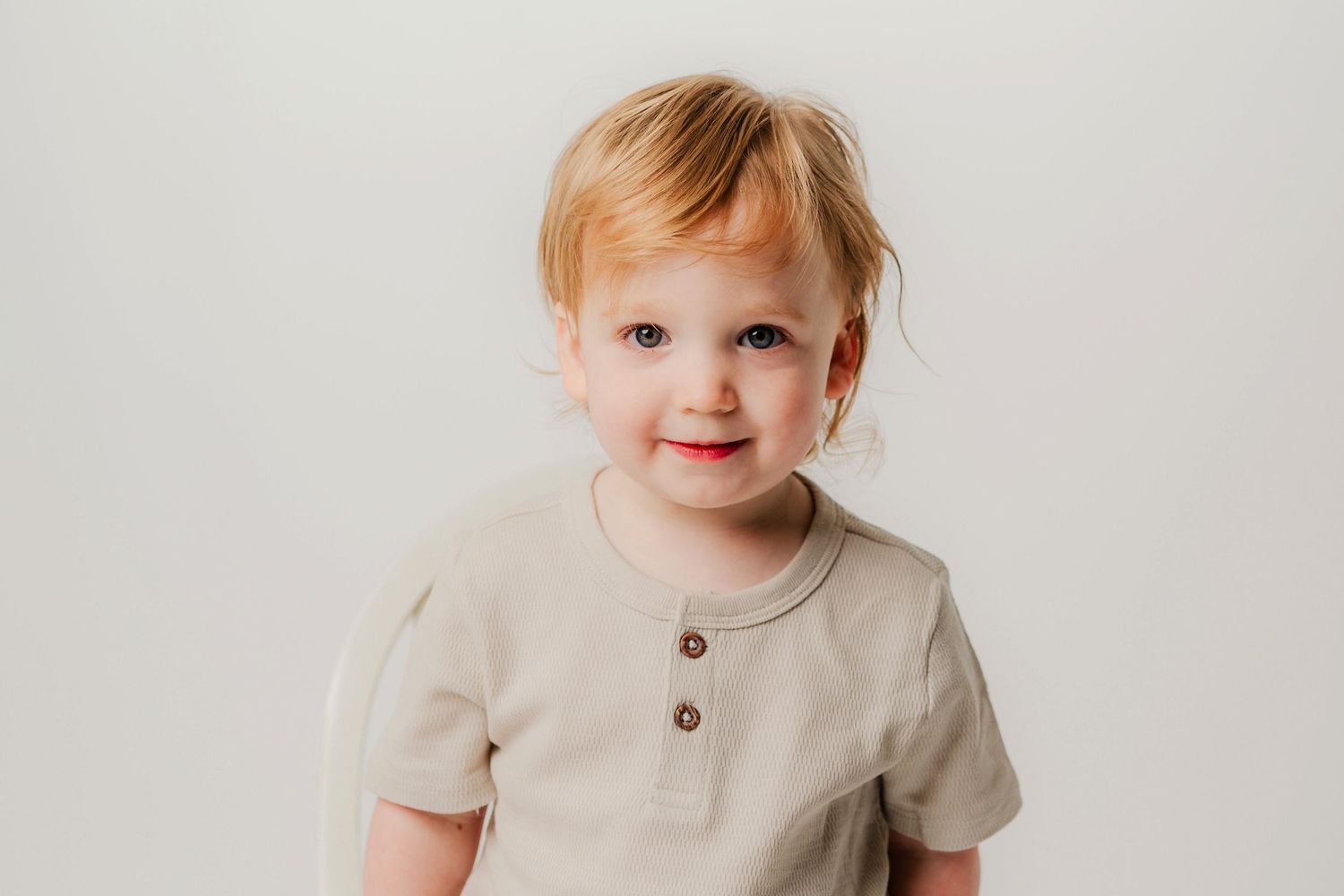 A toddler wearing a light colored henley shirt smiles against a plain light background.