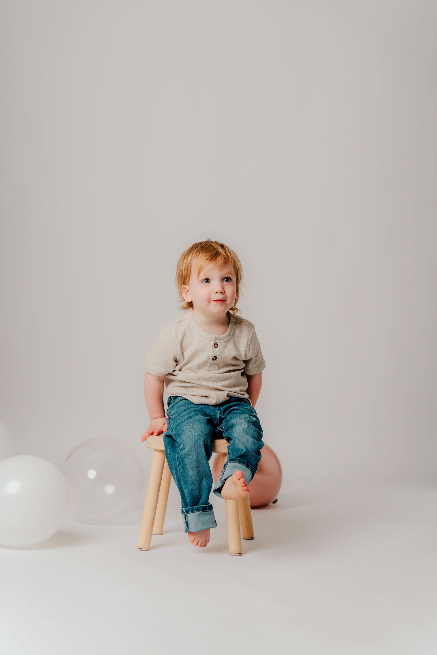 A small child sits barefoot on a wooden stool wearing jeans and a light colored t-shirt against a neutral background.