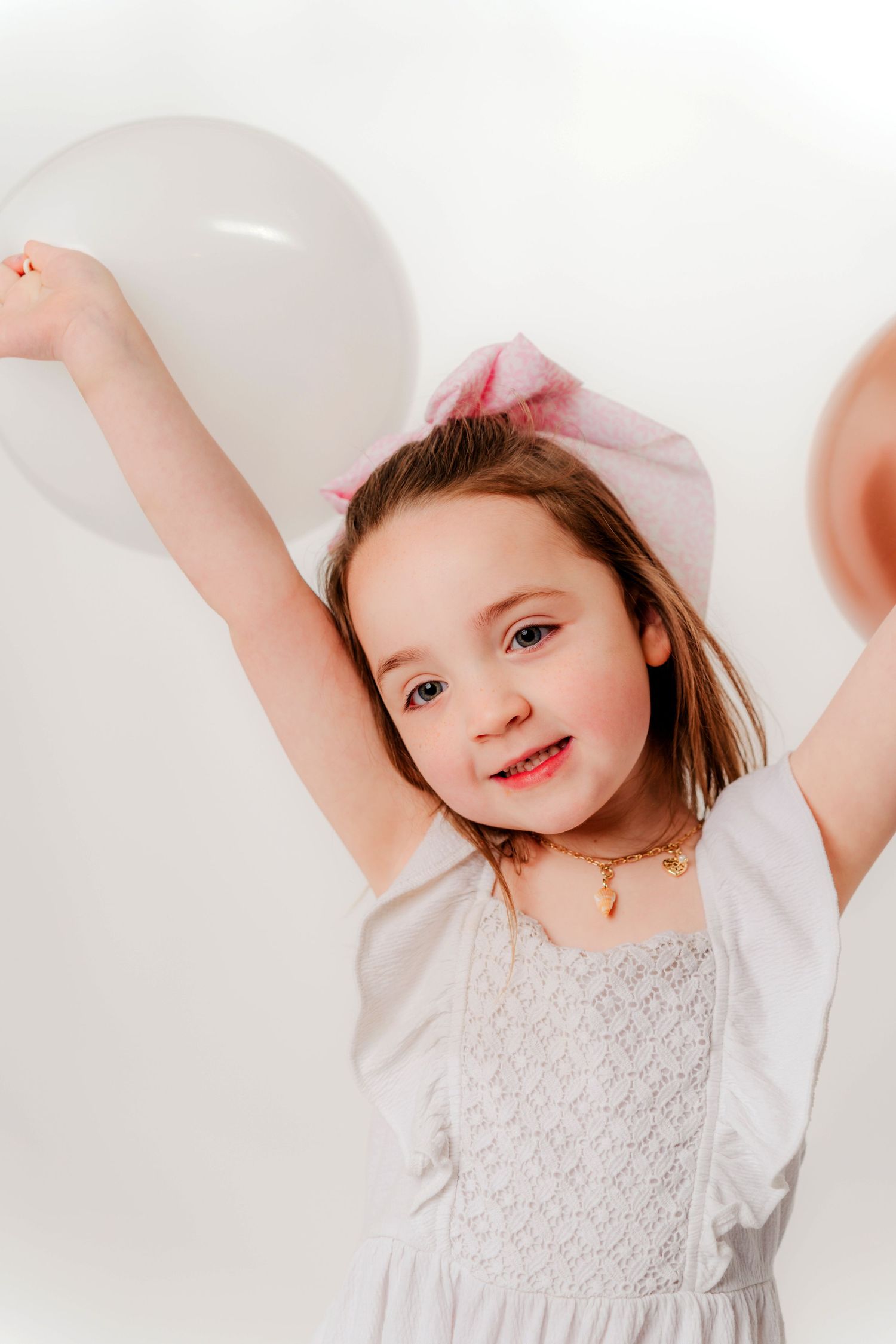 Young child in white dress with pink bow excitedly raises arms while holding white balloons against white background.