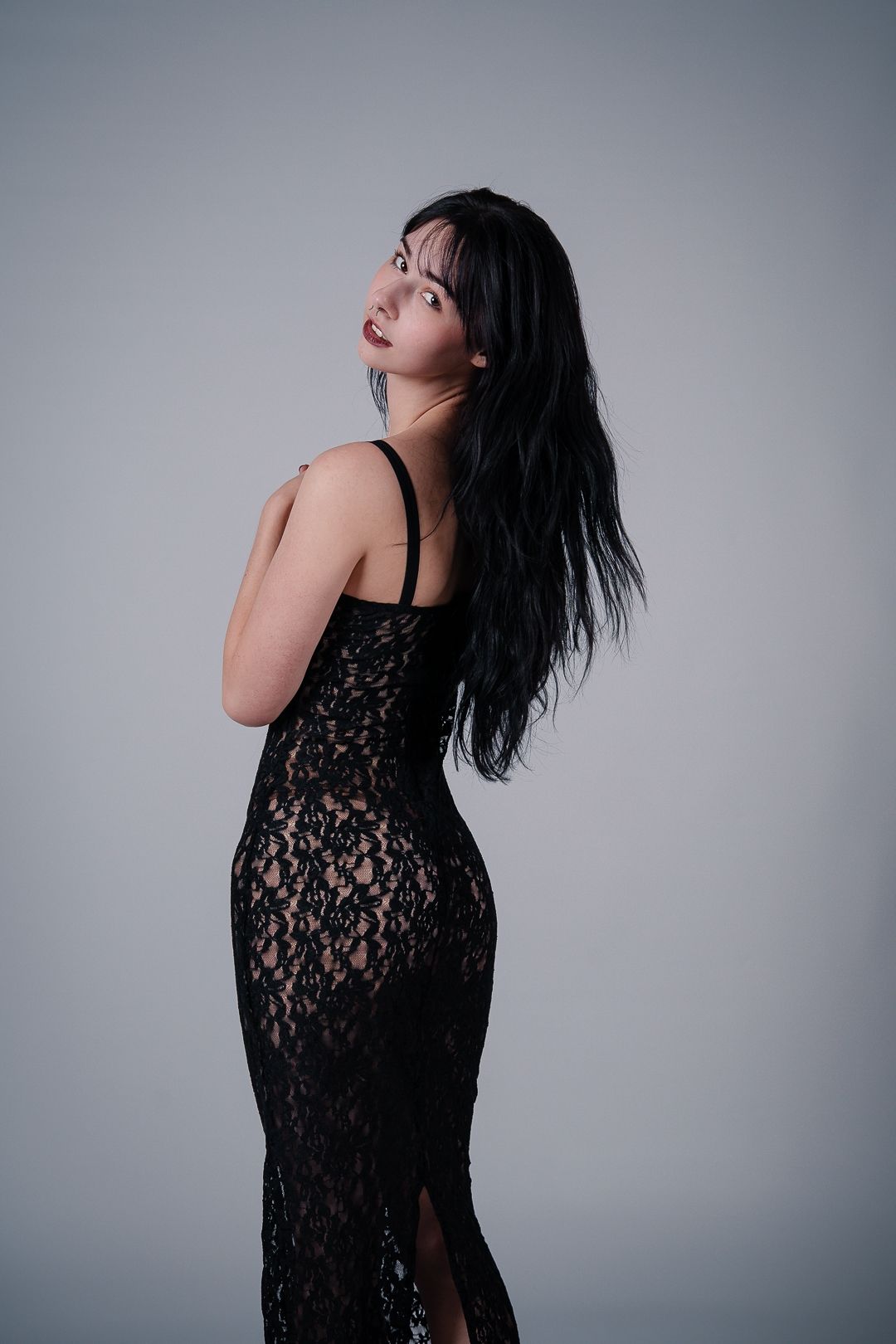 A person in a black lace evening gown poses elegantly against a gray studio backdrop.
