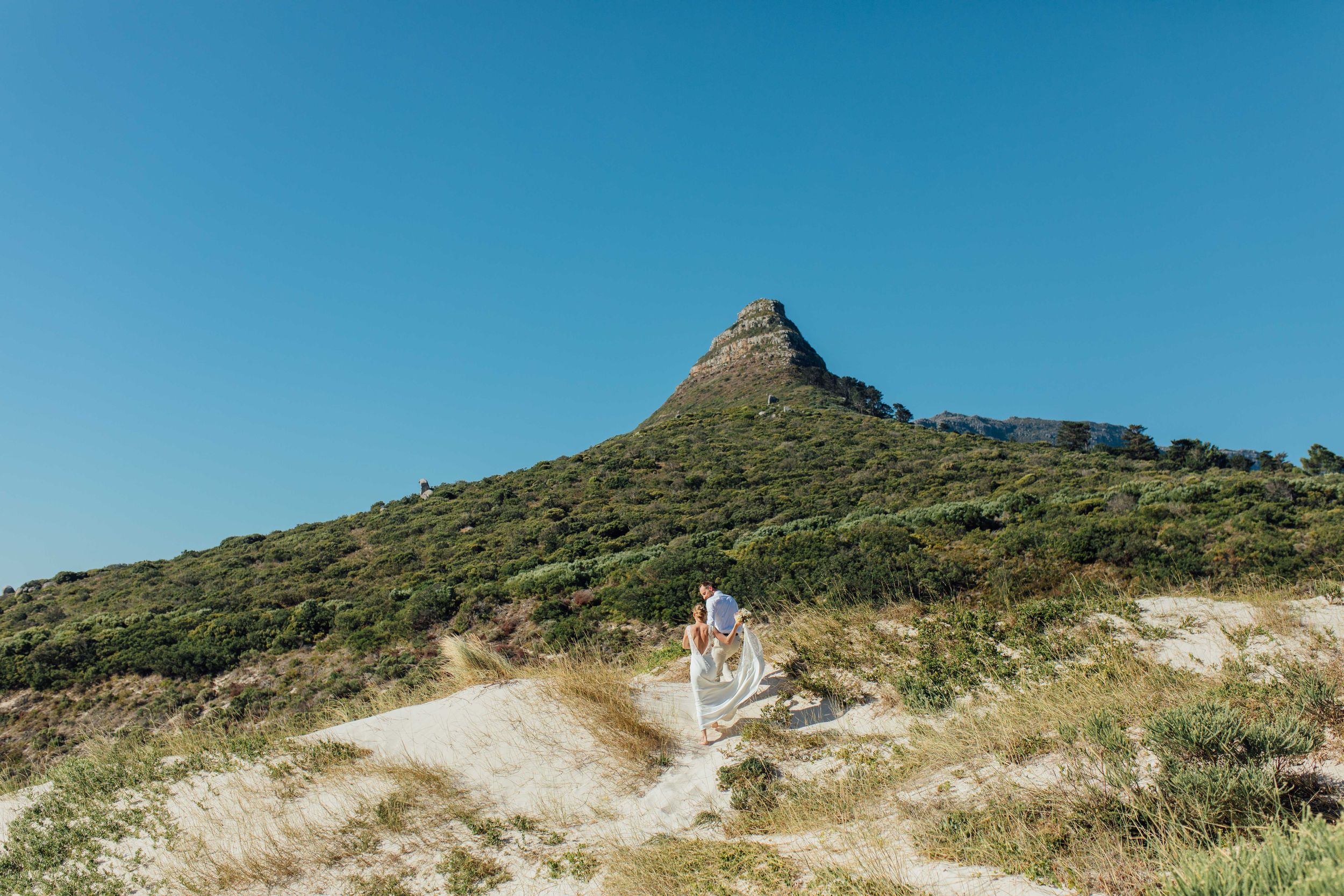 A newlywed couple walks  along a dirt trail towards a distinctive mountain peak on a clear sunny day.