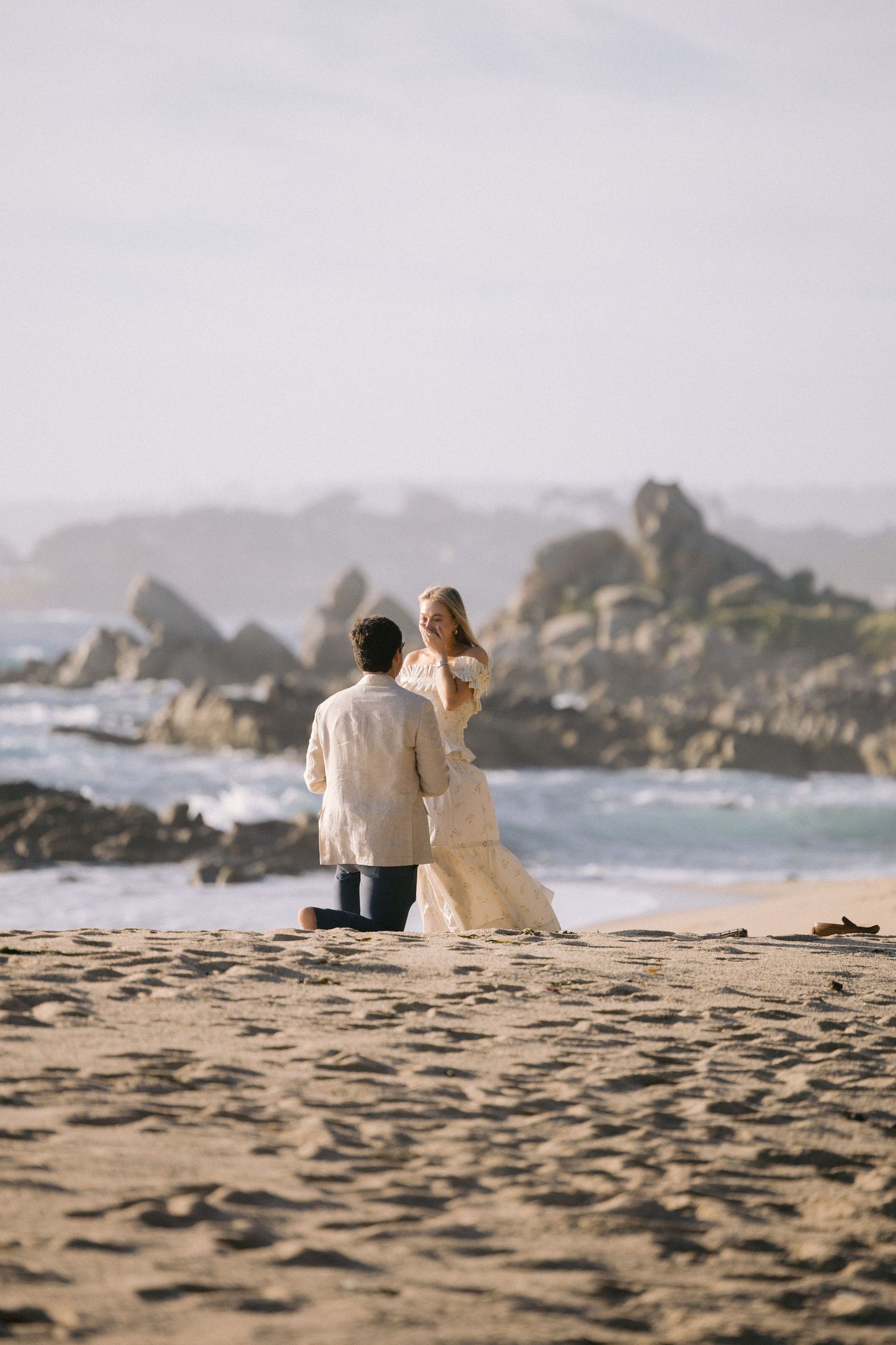 A couple in wedding attire stands on a sandy beach with rocky coastline and ocean waves in the background.