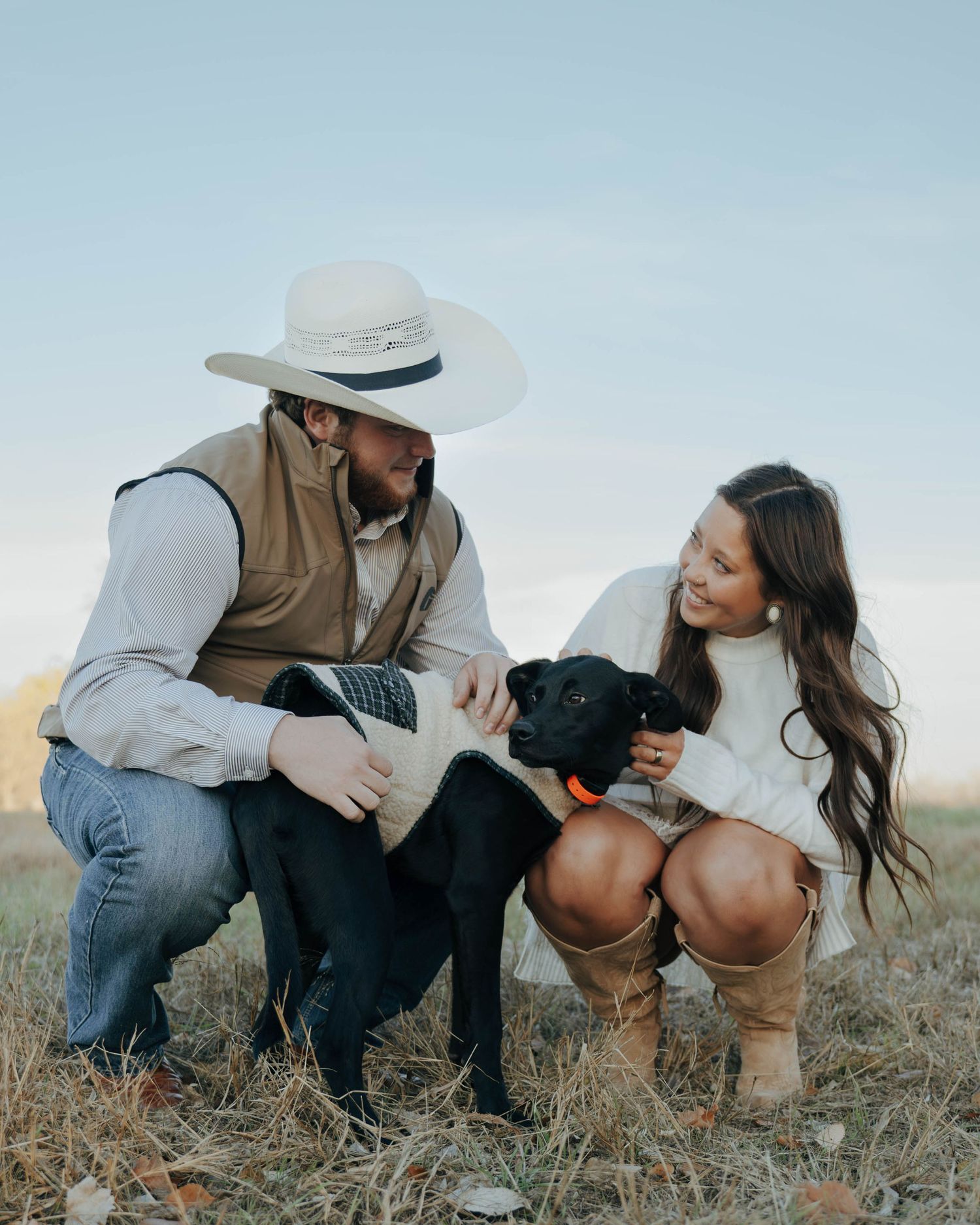 A couple and their black dog enjoy a sweet moment together in a grassy field on a sunny day.