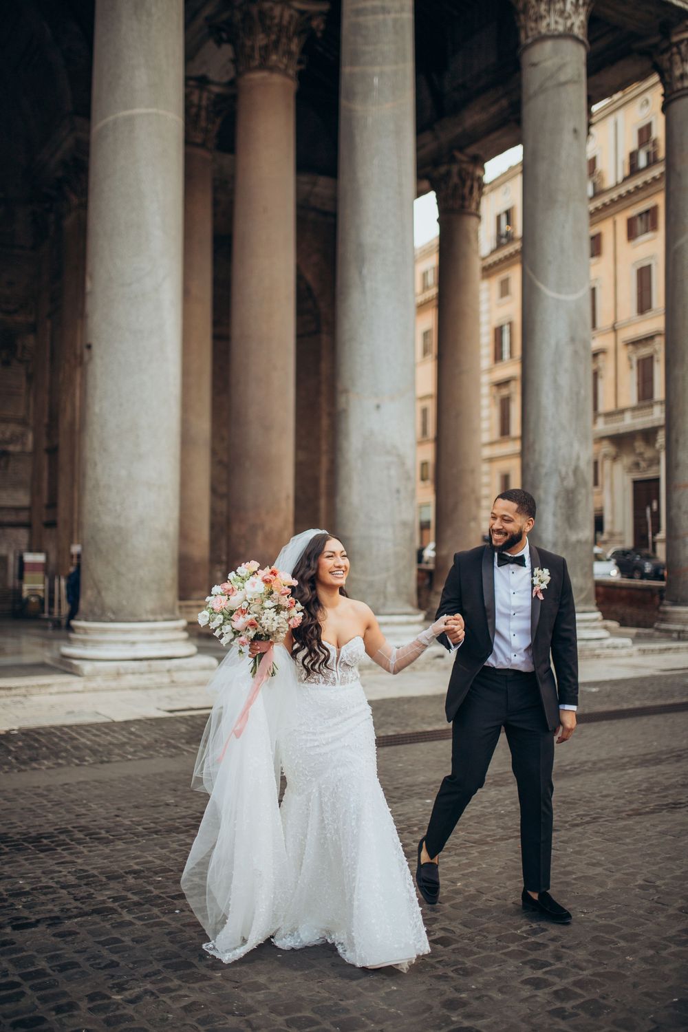 Wedding couple walking hand in hand between ancient Roman columns with a beautiful floral bouquet.