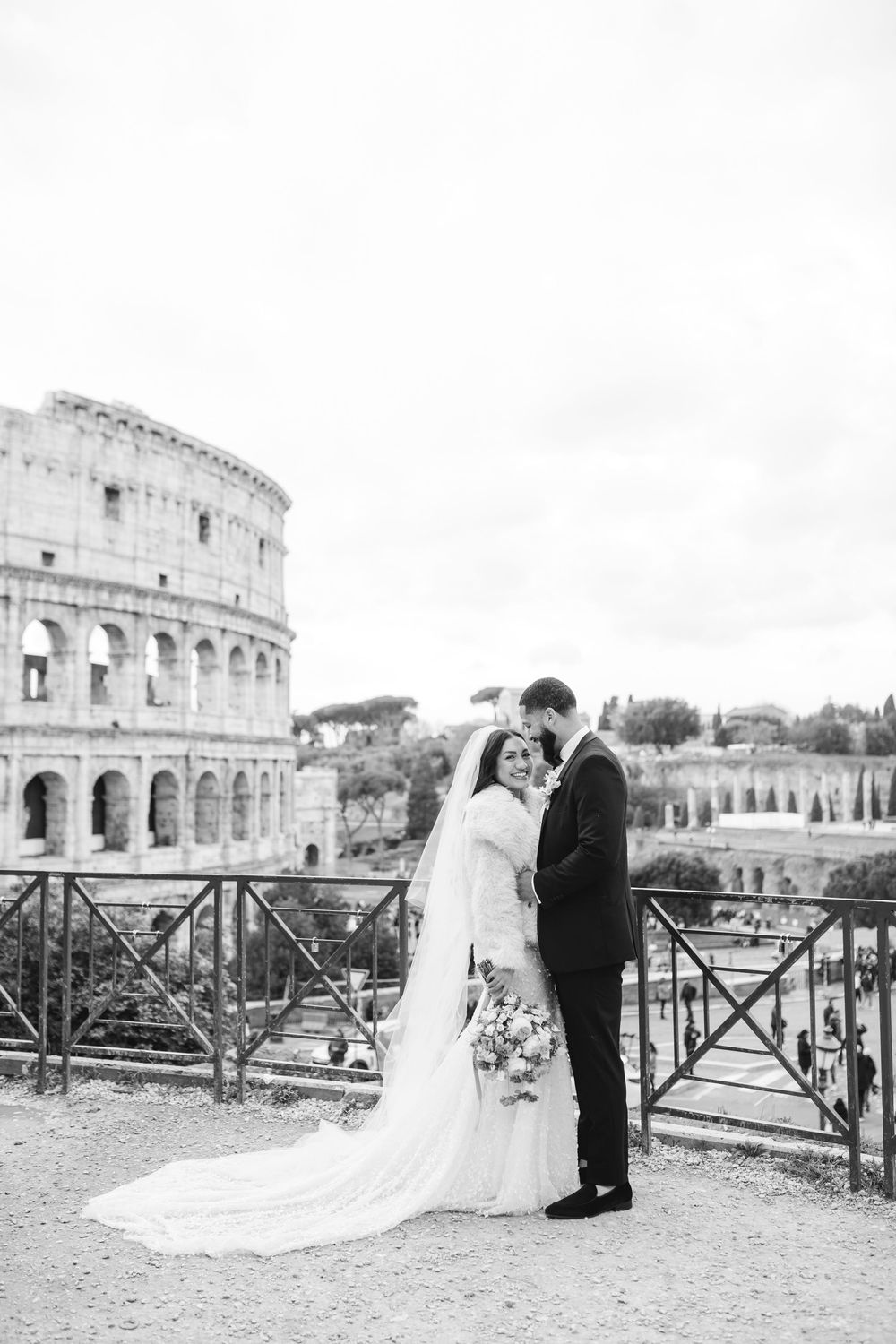 Black and white portrait of newlyweds embracing near historic Colosseum in Rome with elegant bridal attire.
