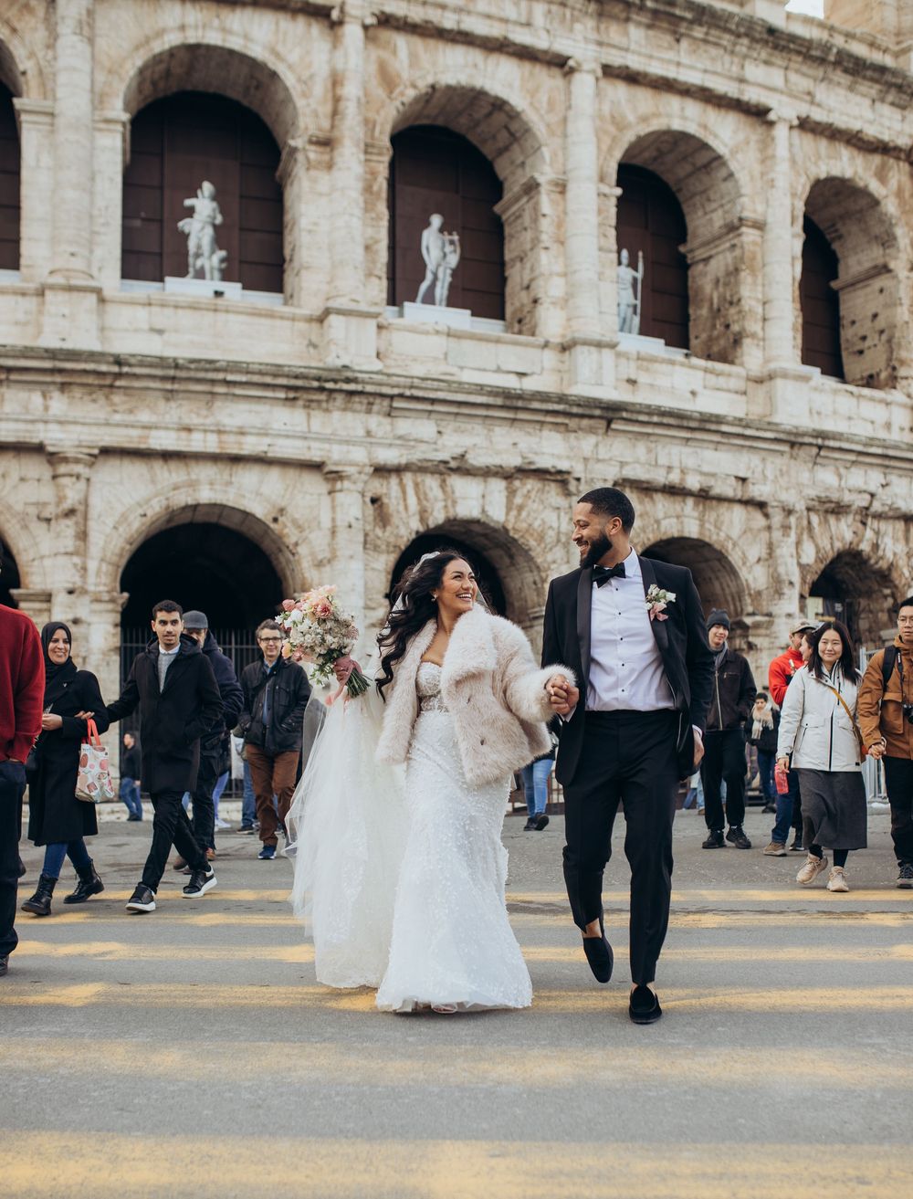 A couple walks in formal attire outside the ancient Colosseum in Rome among tourists.
