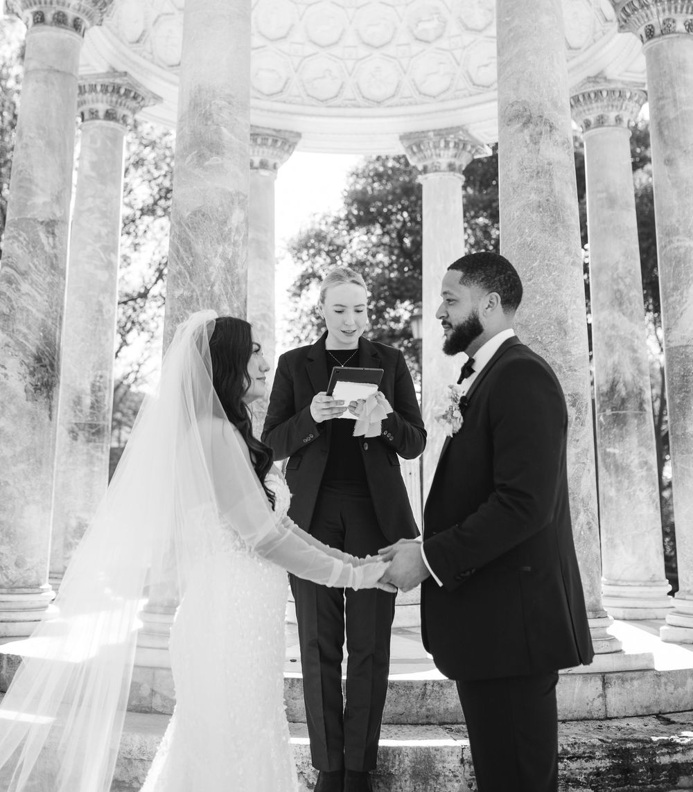 A wedding ceremony captured in black and white under an elegant columned pavilion with ornate architectural details.