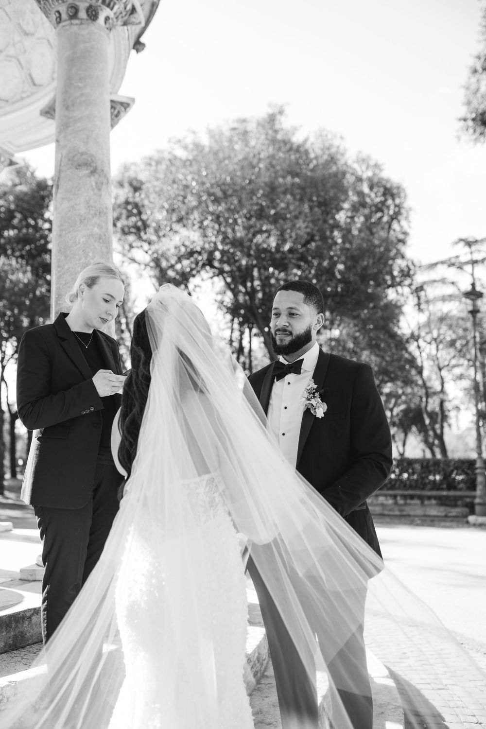 A black and white wedding photo showing a ceremony under a classical stone gazebo with a flowing bridal veil.